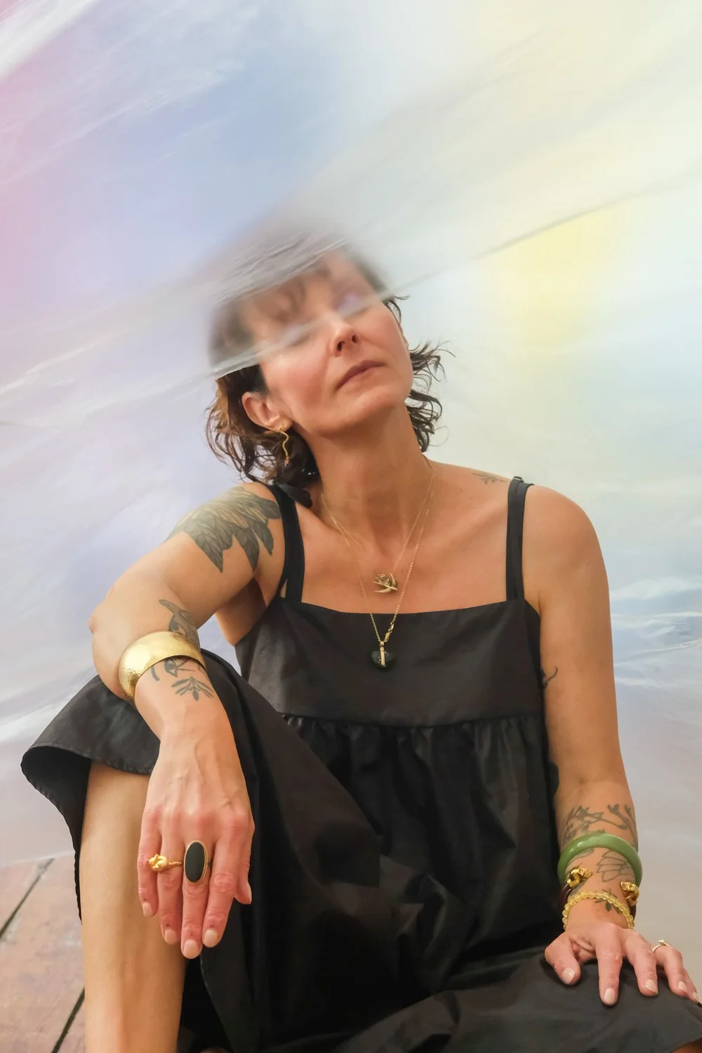 Een vrouw met tattoos en sieraden, gekleed in een zwarte jurk, zittend op de vloer, met een wazige reflectie boven haar hoofd.