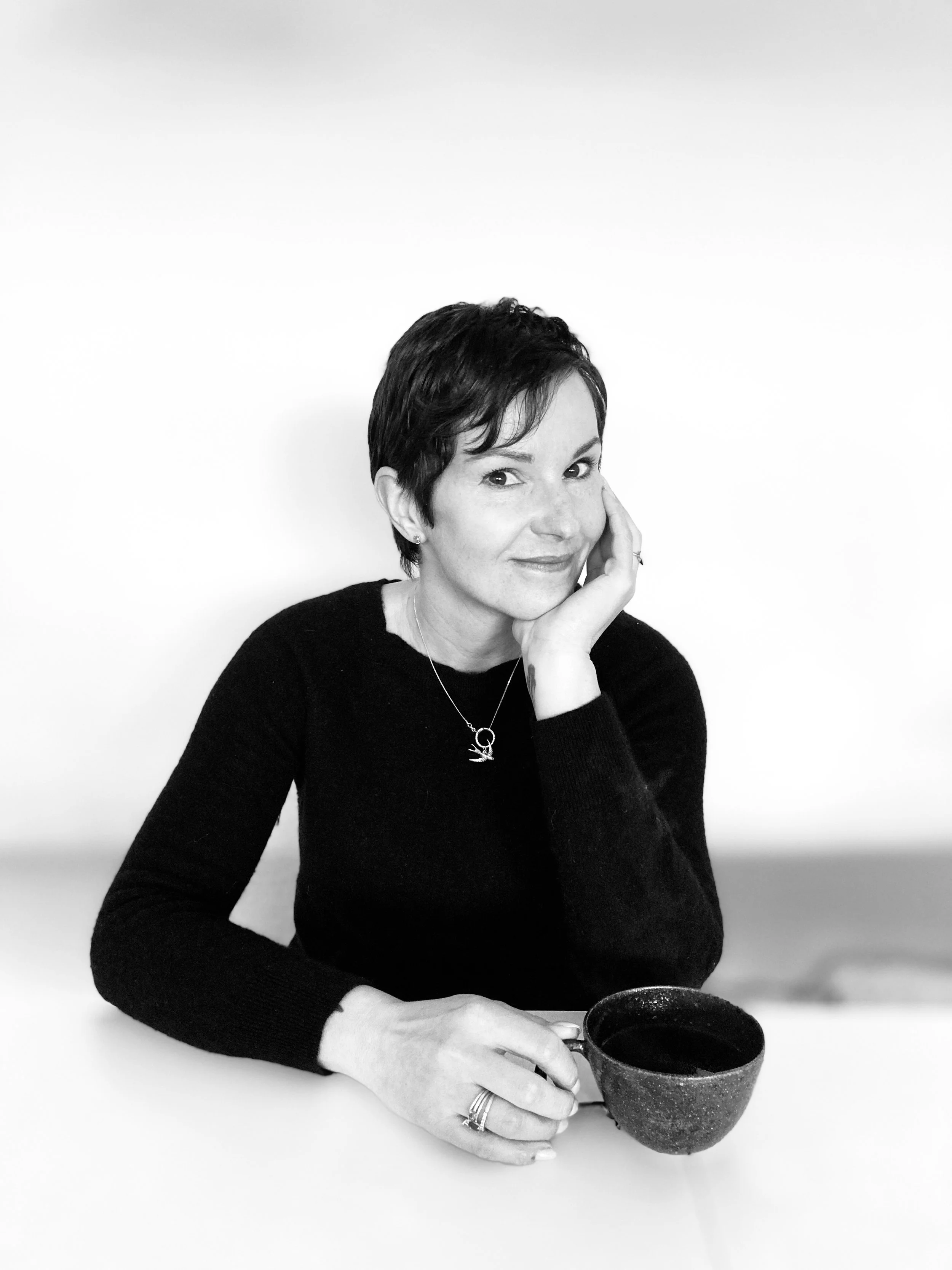 Black and white portrait of a person with short hair wearing a black sweater, holding a cup and leaning on a table.