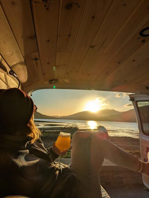 Person relaxing in a camper van at sunset, overlooking a lake and mountains.