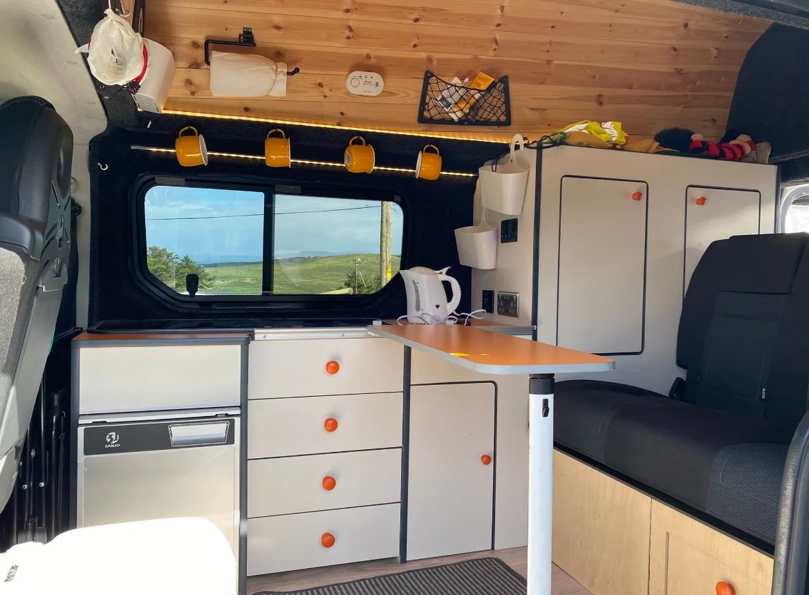 Interior view of a camper van with a window showing green hills and blue sky, white cabinets with orange knobs, a small fridge, a black couch, a foldable table, a kettle, hanging mugs, and various small storage items.