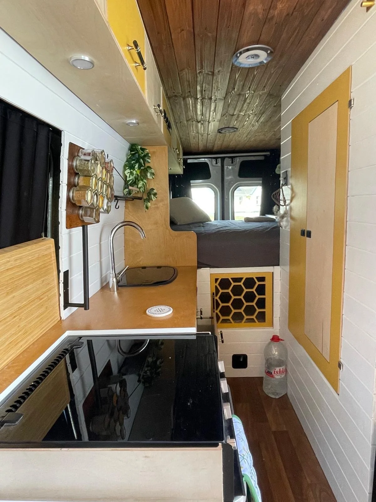 Interior view of a compact kitchen and bedroom area inside a camper van, with a small sink, stove, spice jars, a potted plant, and a bed visible through a doorway.