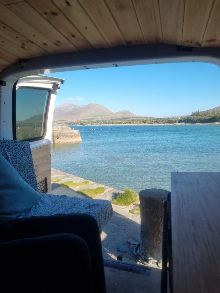View from inside a van showing a body of water with mountains in the background, a concrete pier, and a clear blue sky.