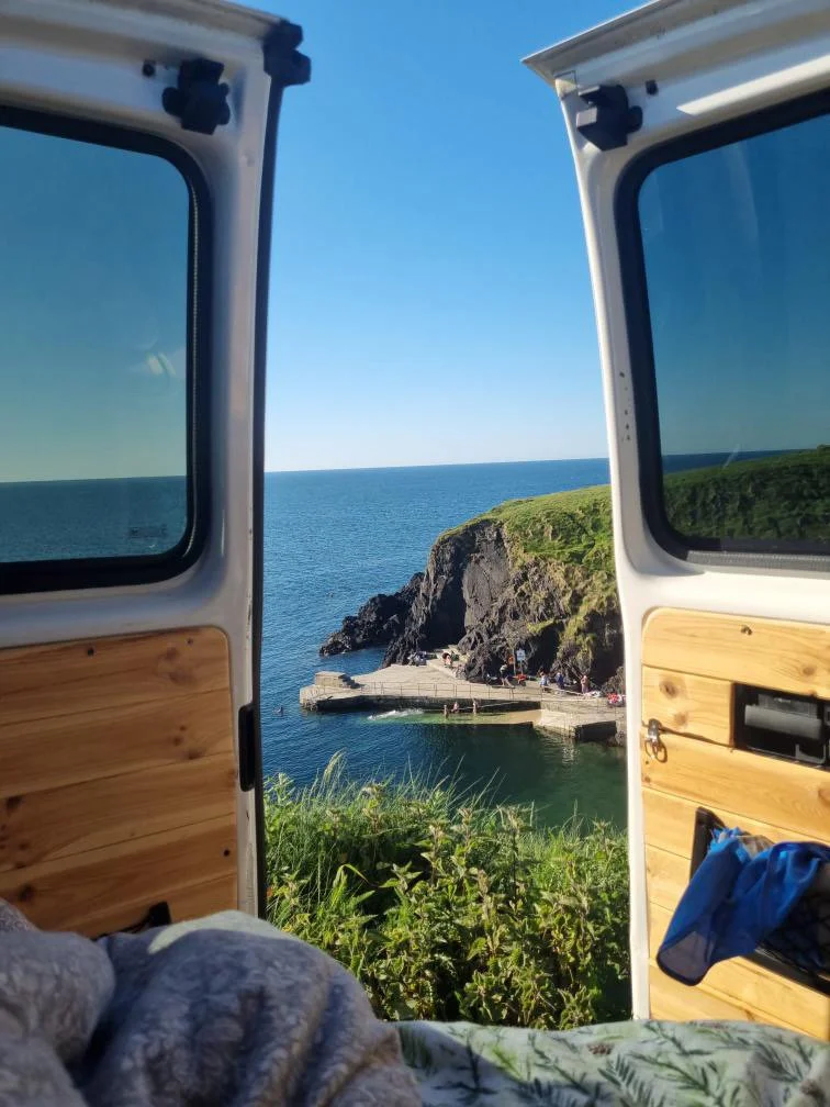 View of a rocky coastline and ocean through open camper van doors, with a small pier and green hillside in the background.