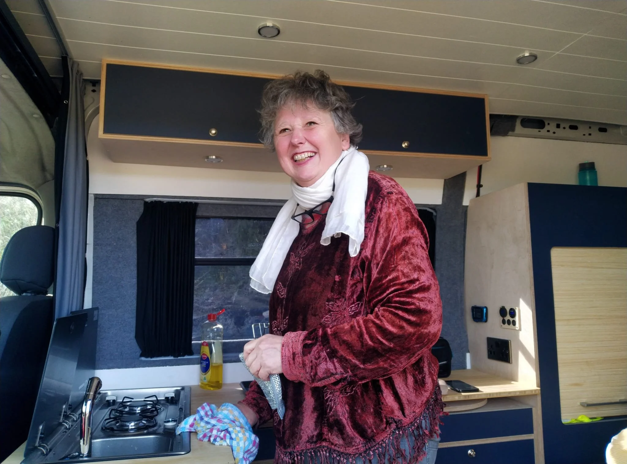Smiling elderly woman with short curly hair, wearing a white scarf and a maroon velvet jacket, inside a tiny home, holding cleaning cloths near a small gas stove.