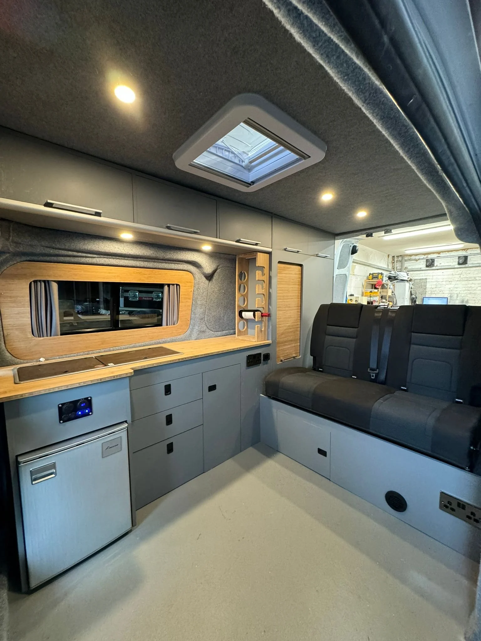 Interior of a converted van with gray cabinets, a wooden countertop, a small fridge, a window, and a black seating area with seat belts.