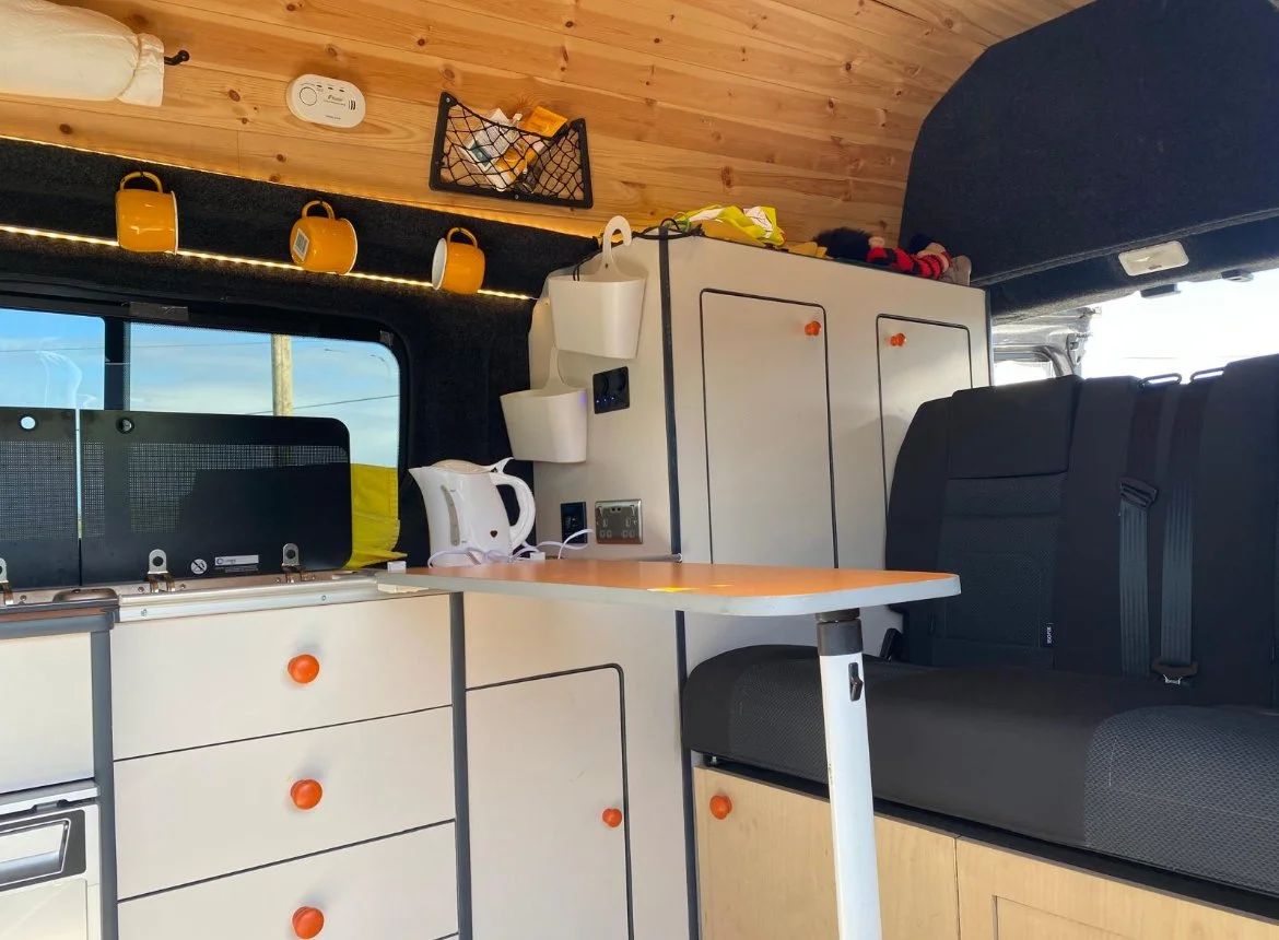Interior of a camper van with white cabinetry, orange knobs, a black sofa, a small table, a window, and kitchen items including a kettle and mugs.