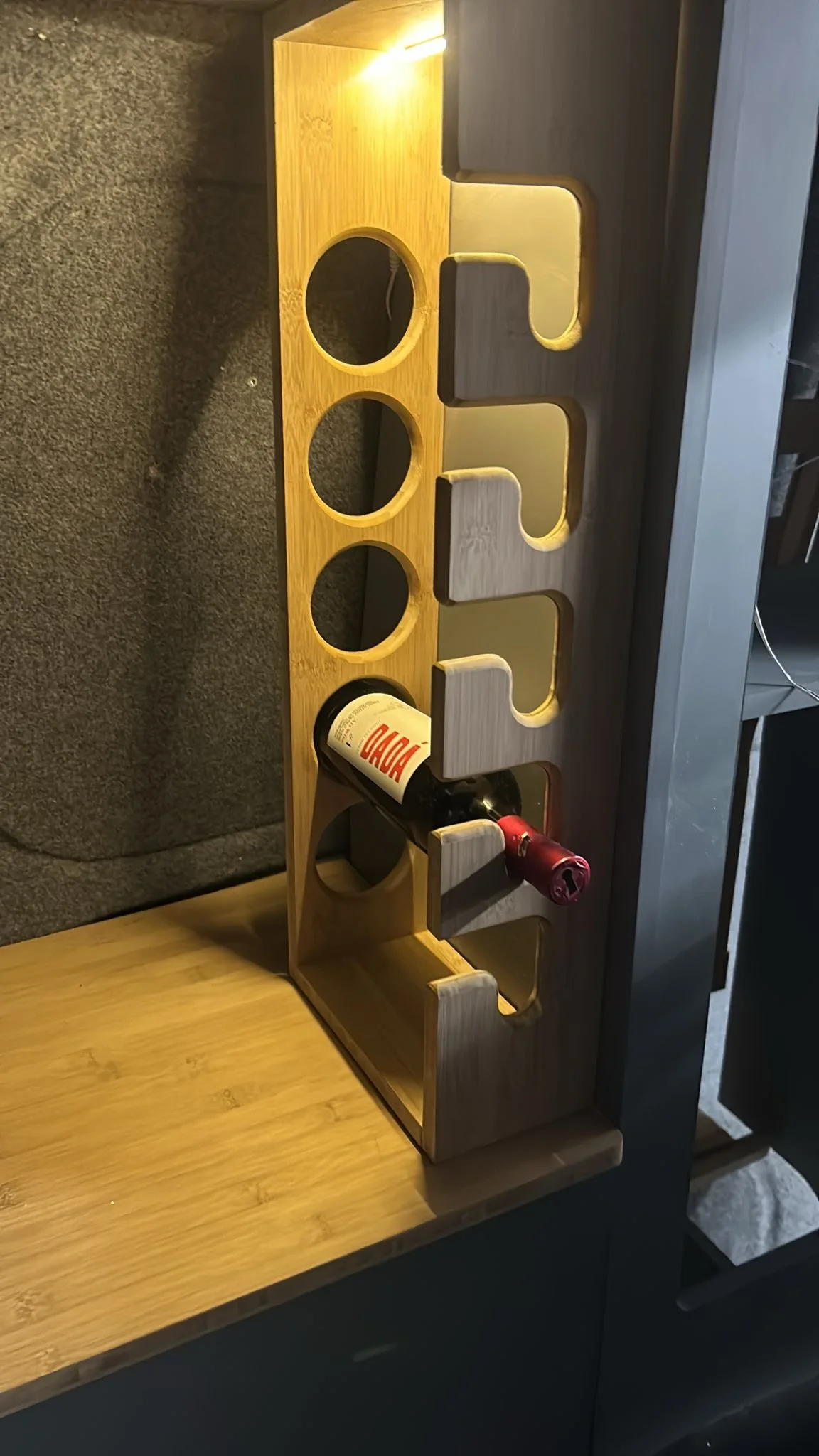 A wooden wine rack with circular holes designed to hold wine bottles horizontally. Two wine bottles are placed in the rack, one with a red capsule and one with a black capsule. The rack is on a wooden surface next to a gray sofa.