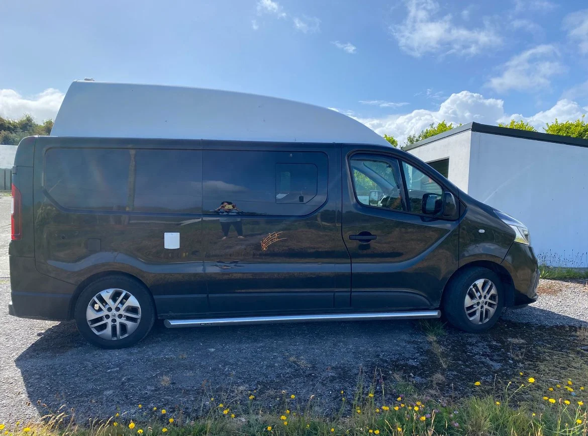 Black camper van parked outside on a sunny day, with a white building and greenery in the background.