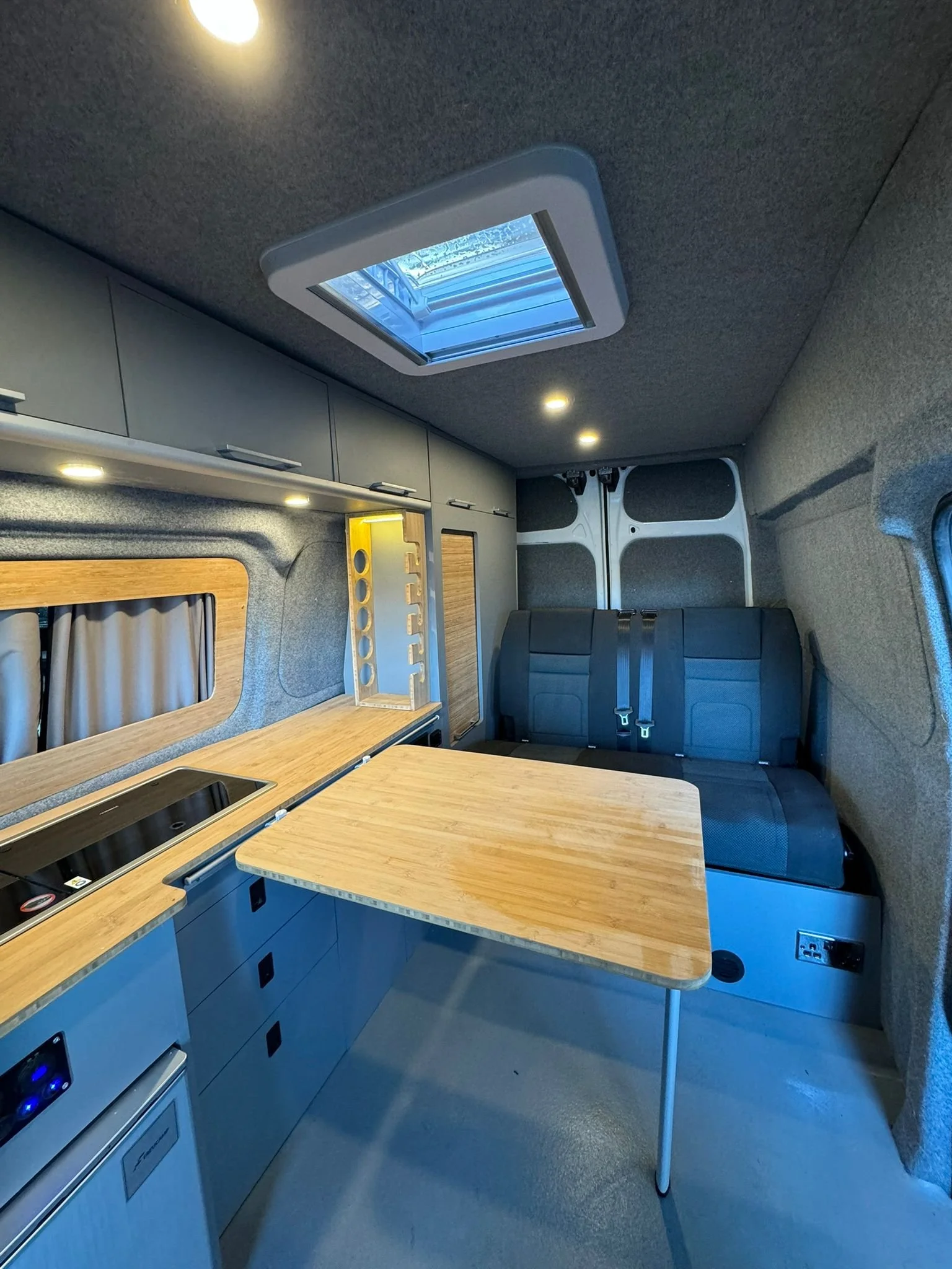 Interior of a modern camper van with a wooden table, kitchen area, storage cabinets, a skylight, and a seating area with black seats and seatbelts.