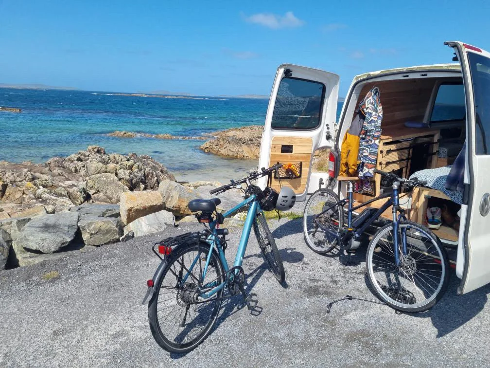 Two bicycles parked outside a camper van near a rocky beach with the ocean and a blue sky in the background.