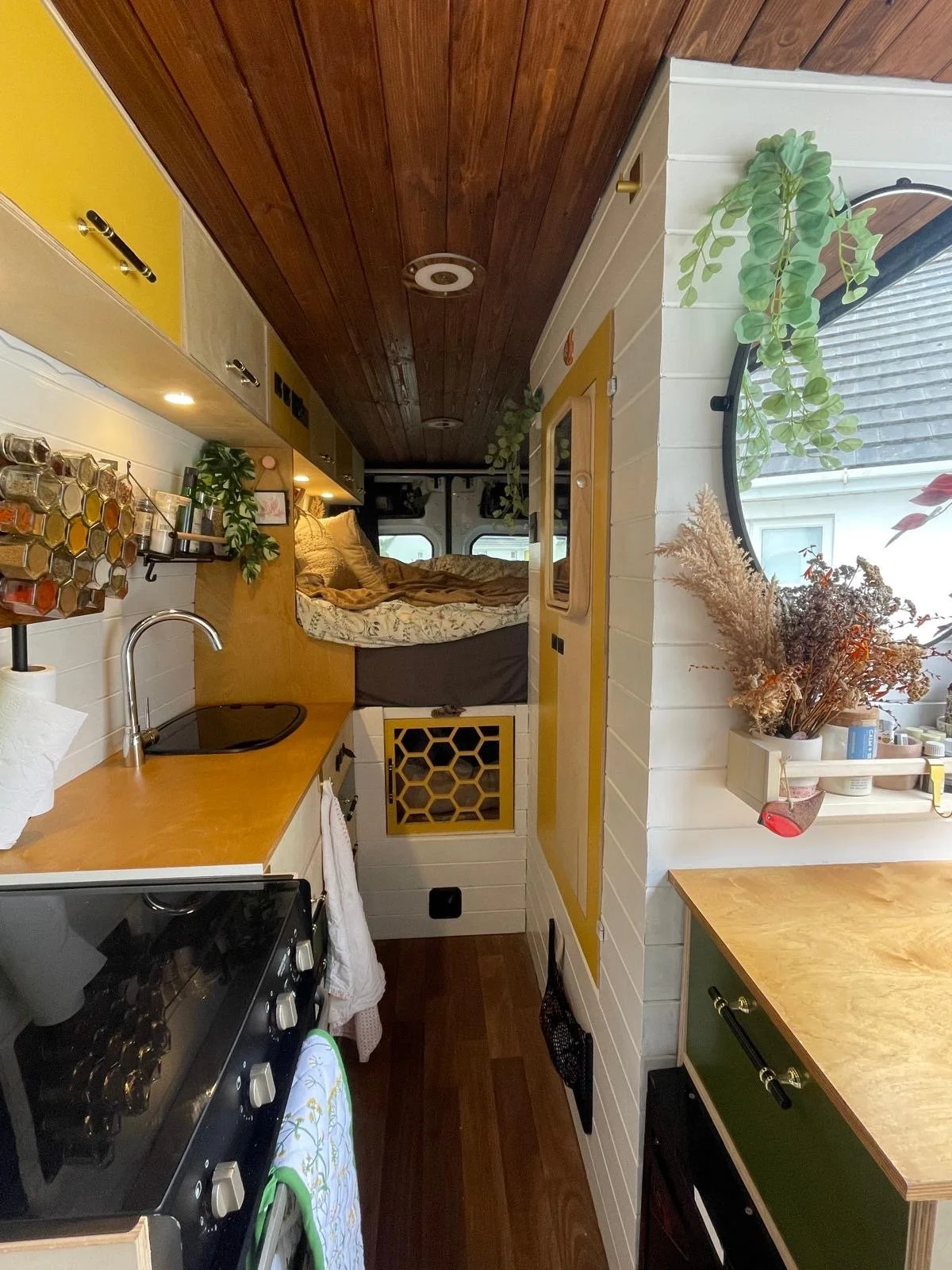 Interior of a tiny house on wheels with a small kitchen, bed area, and round window with plants.
