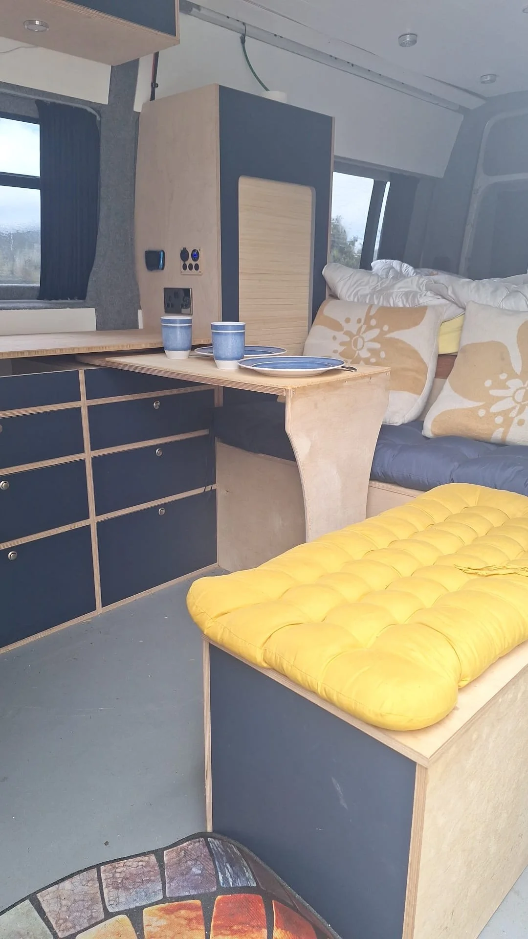 Interior of a camper van with a small table set for two, navy blue cabinets, a yellow cushioned bench, and a bed with white and beige patterned pillows near a window.