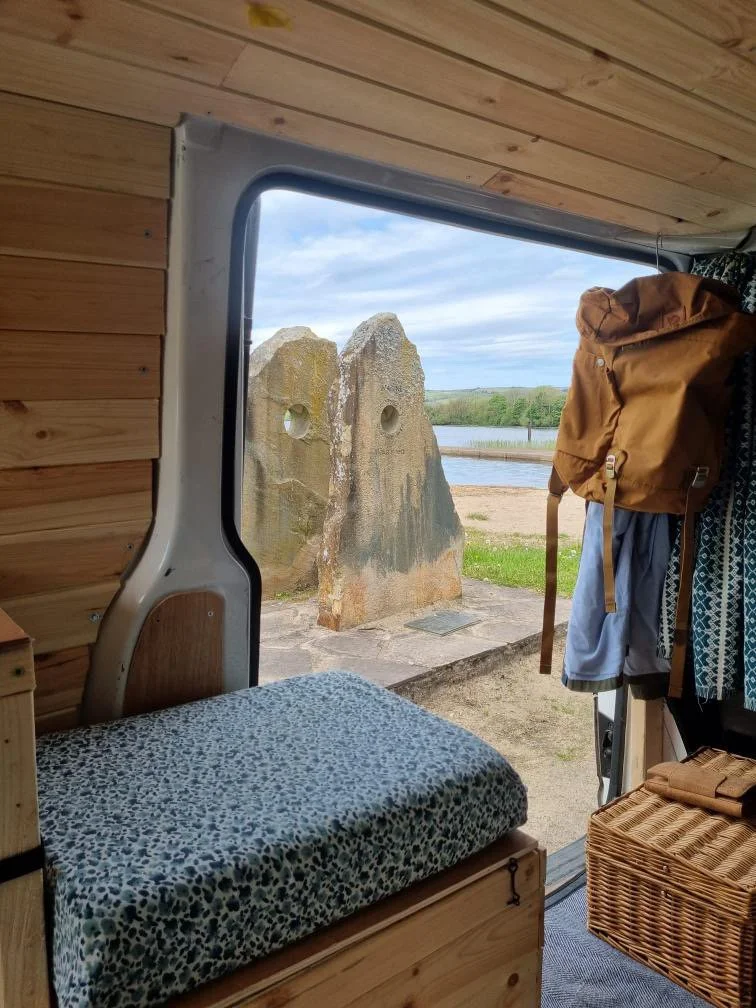 View through a camper van window showing two large rocks with holes, a body of water, and a partly cloudy sky.