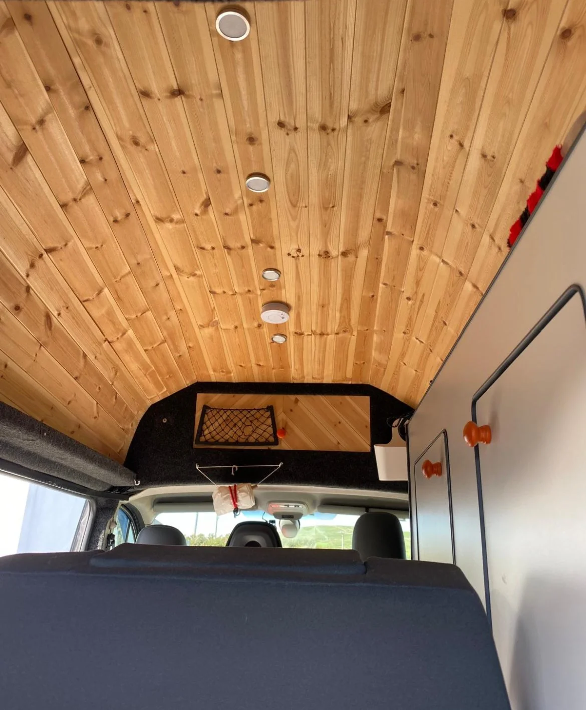 Interior of a camper van with a wood-paneled ceiling, black storage compartments, a mounted net, and a view of the front seats and dashboard through the windshield.