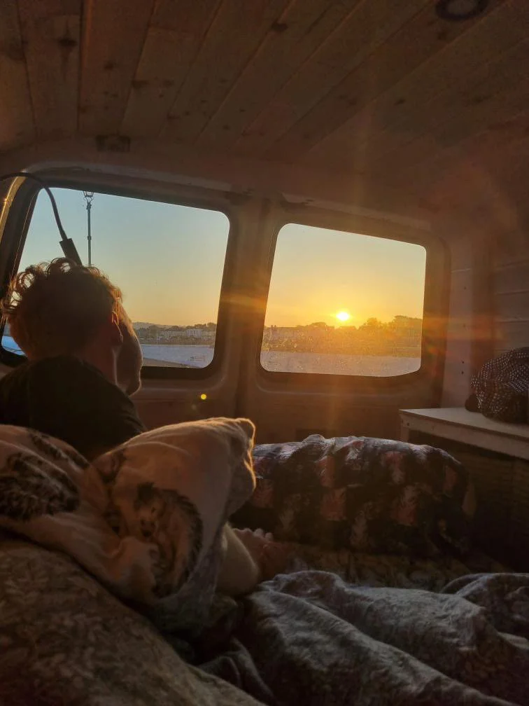 Person relaxing inside a camper van, looking out the window at a sunset over water with a city skyline in the background.