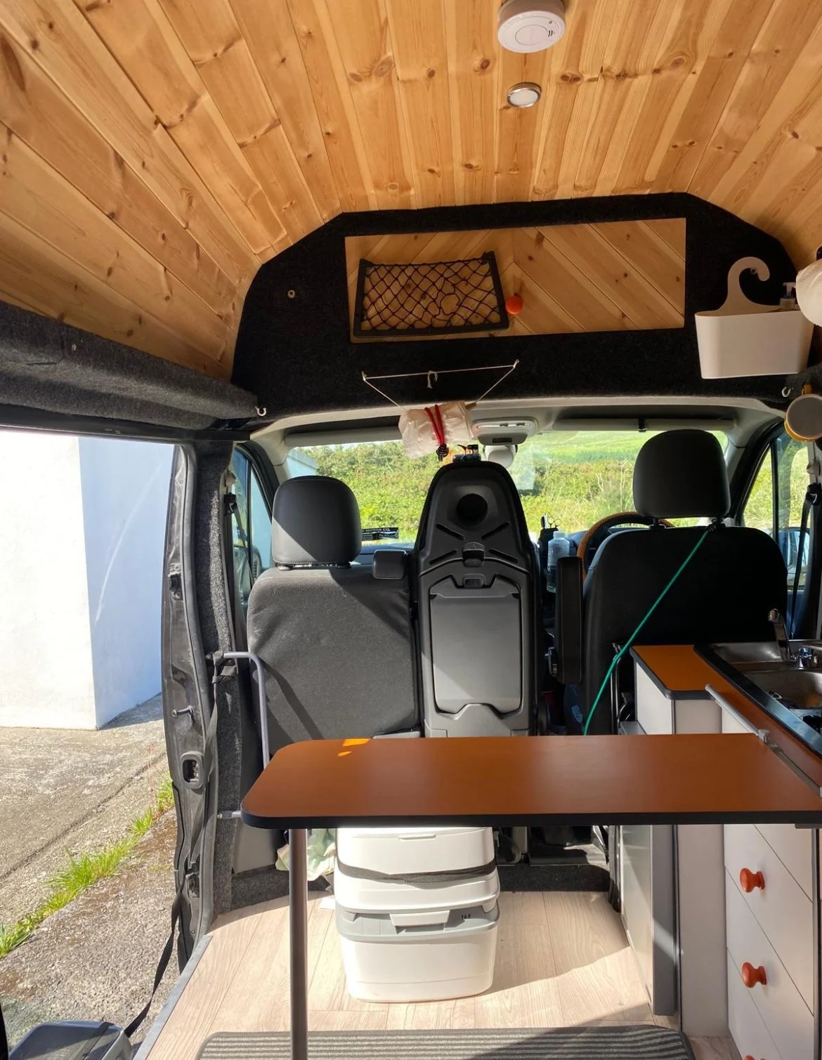 Interior of a camper van with wood-paneled ceiling, a mirror, net storage, and a small sink, with view of green outdoors through the front window.