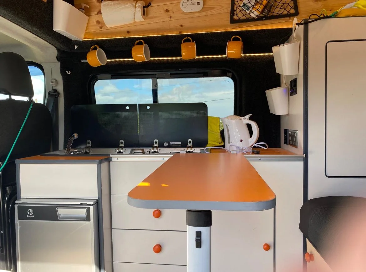 Compact van interior kitchen with small sink, mini fridge, orange countertop, electric kettle, and hanging mugs, with window showing cloudy sky outside.