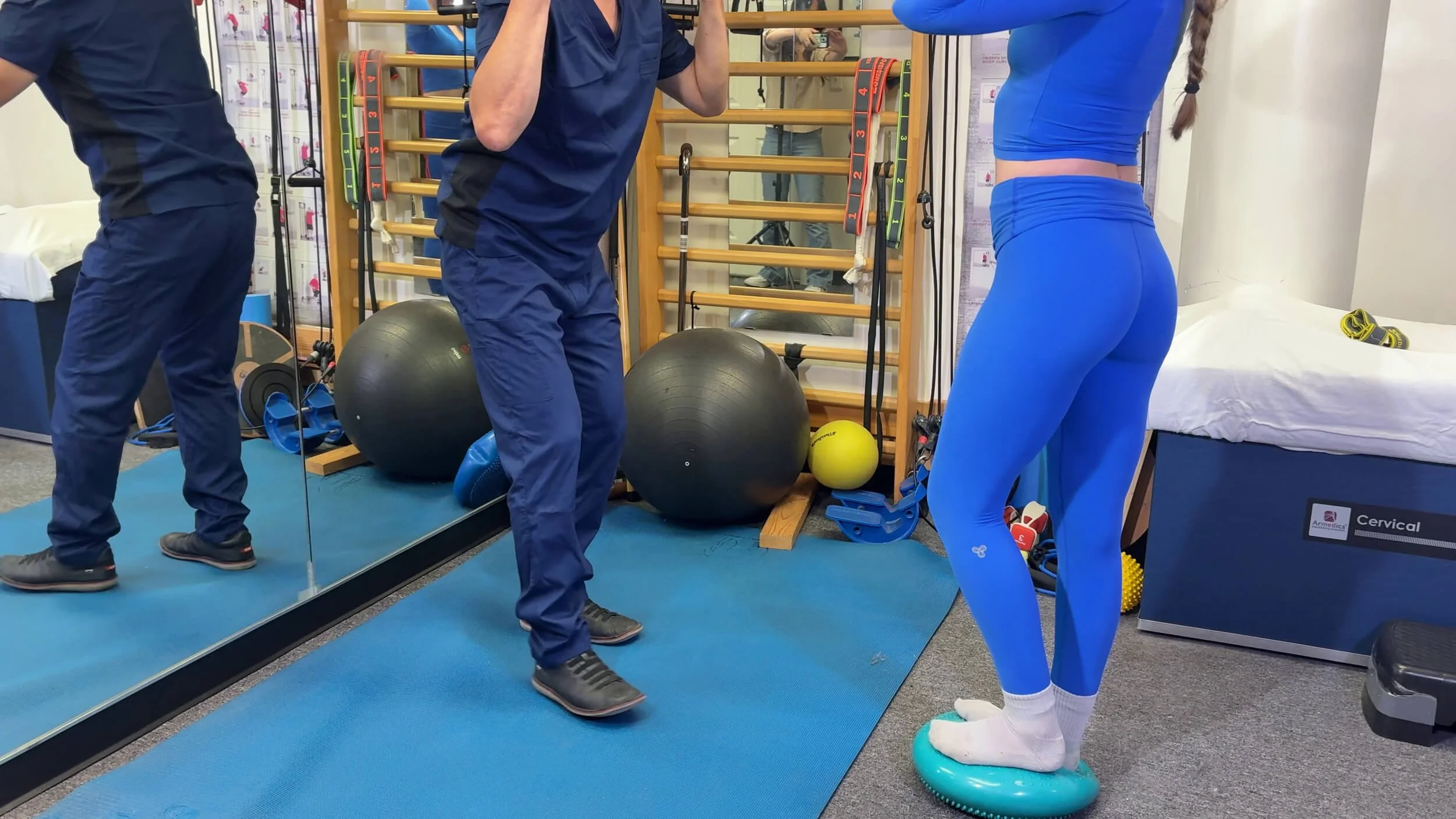 A person in blue athletic clothing stands on a balance disk while a trainer directs the exercise in a fitness room. The room has fitness equipment including stability balls, resistance bands, and a wooden wall with parallel bars.