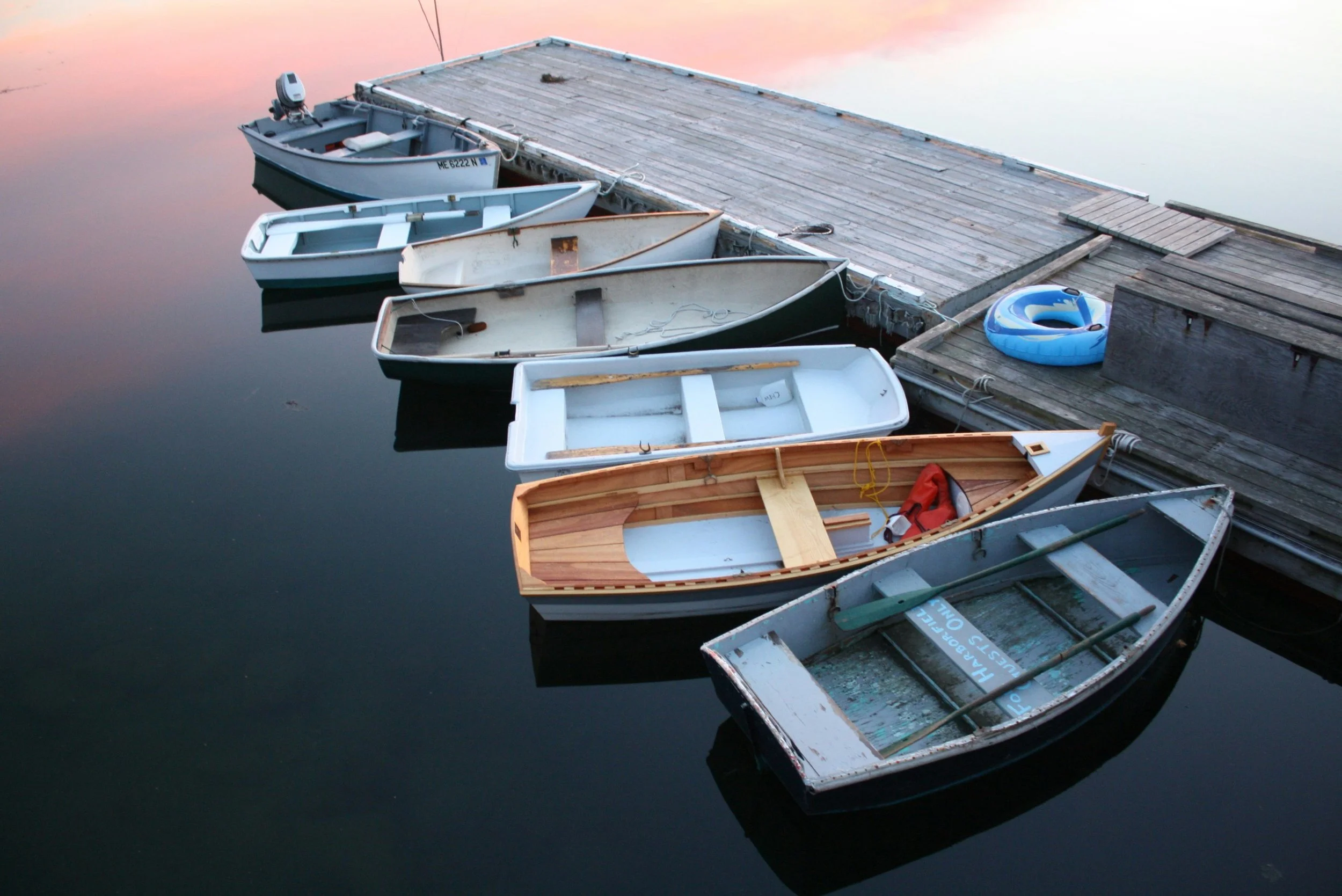 Several small boats docked along a wooden pier on calm water during sunset or sunrise, with one boat featuring a wooden interior and others in various states of use.