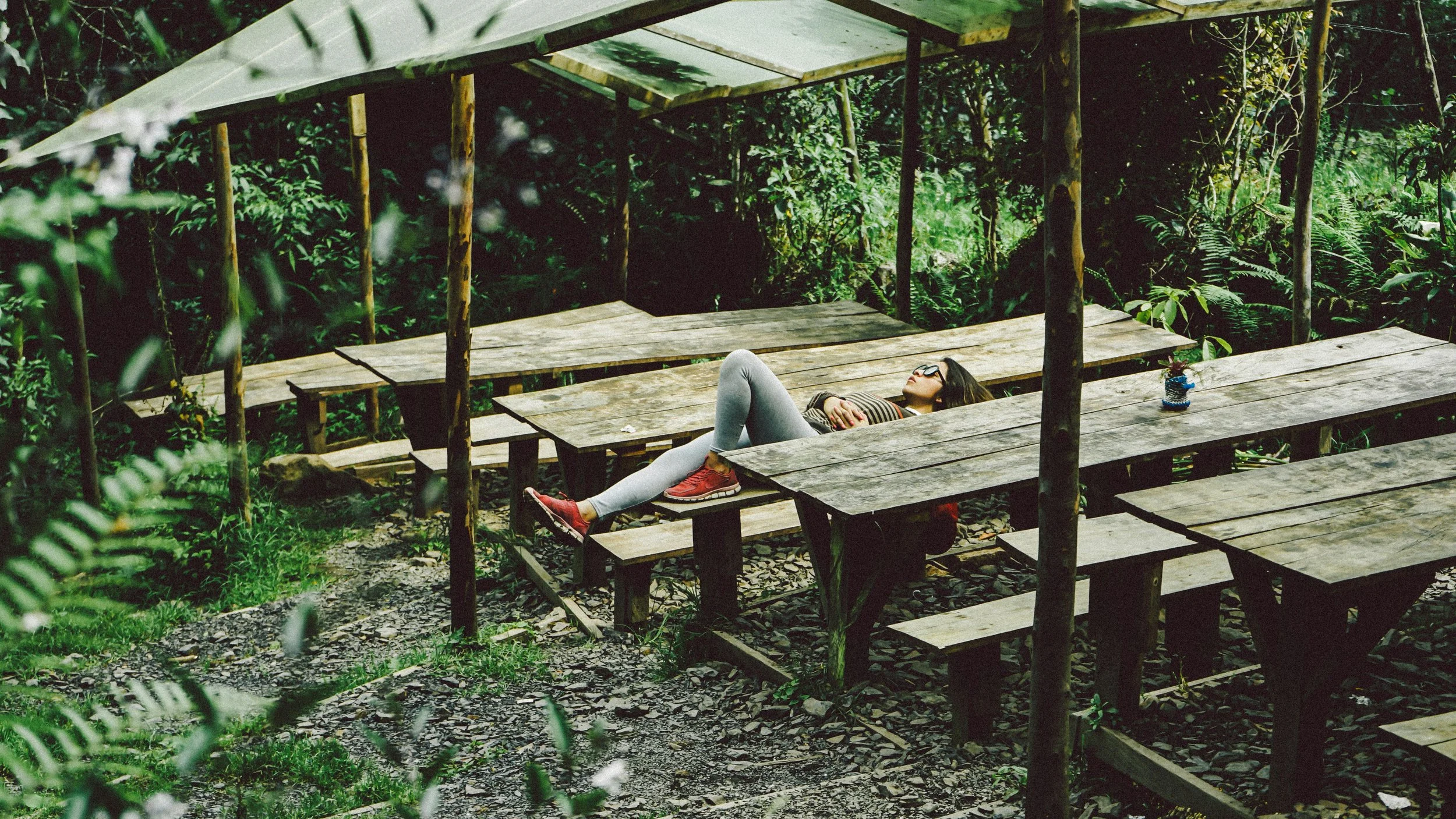 A woman lying on a wooden picnic table in a green forest, resting with her eyes closed, wearing glasses, a striped shirt, gray leggings, and red sneakers.
