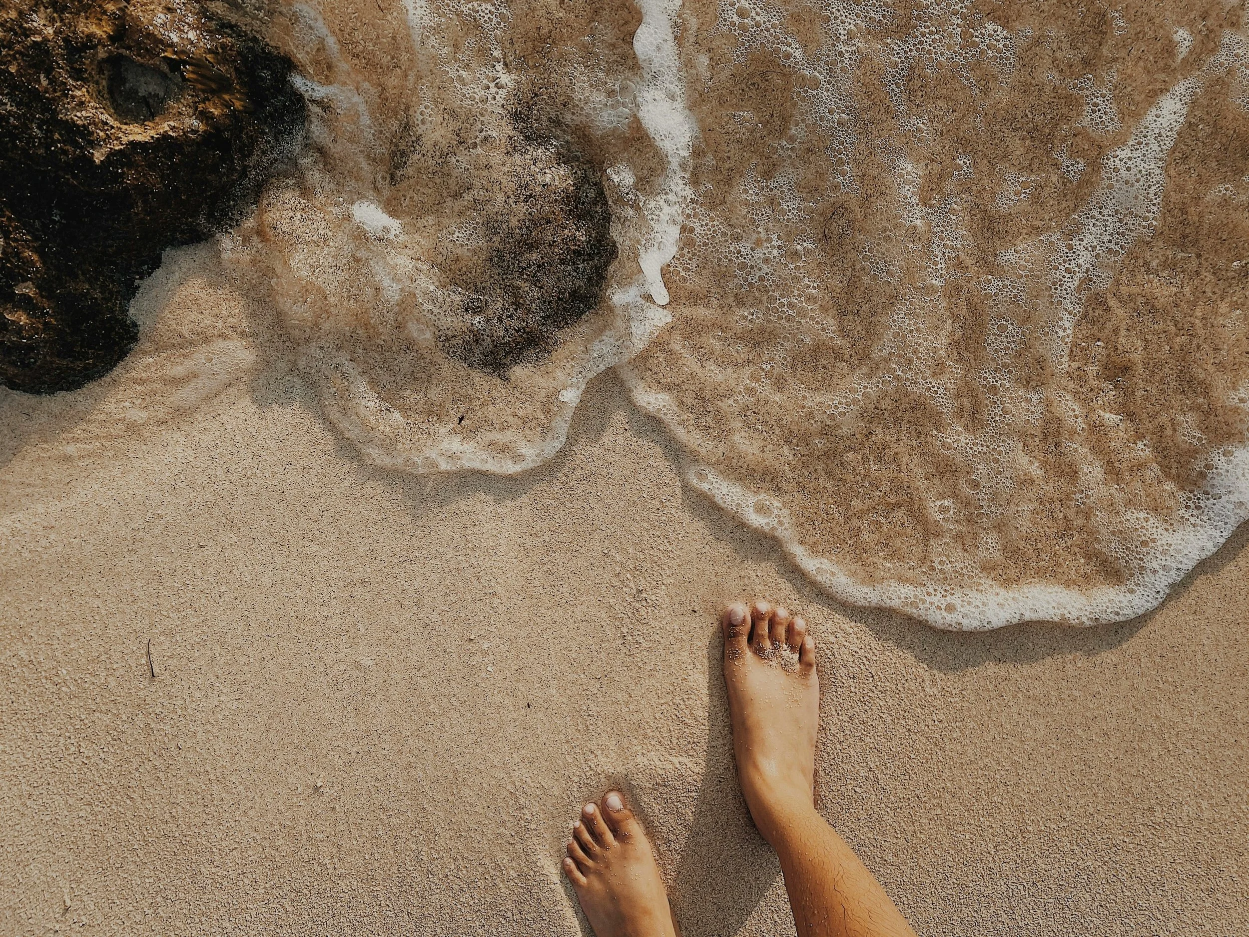 A person's feet on sandy beach with foamy ocean waves approaching.