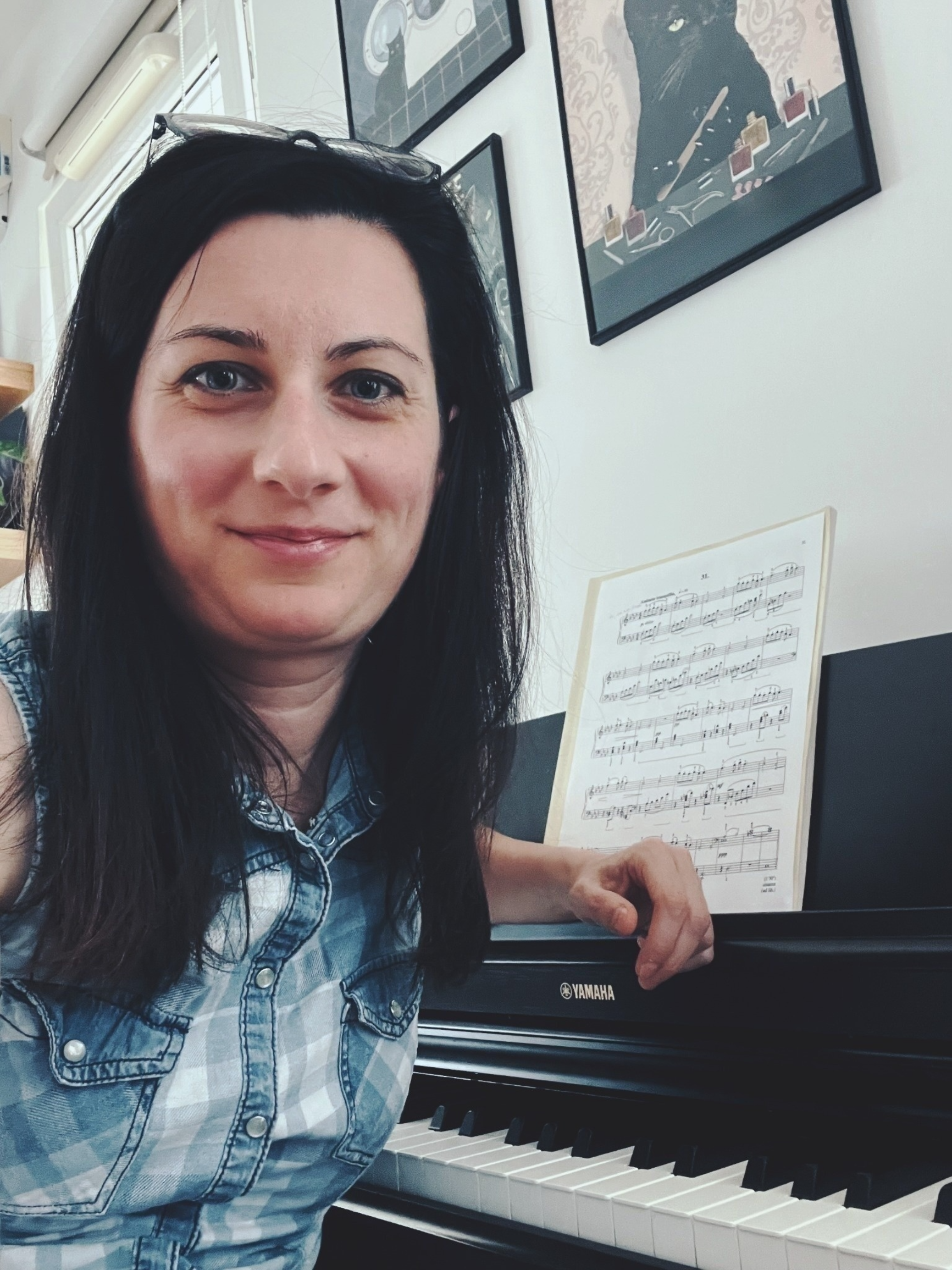 A woman with dark hair smiling at the camera, sitting at a Yamaha piano with sheet music, in a room decorated with framed artwork on the wall.