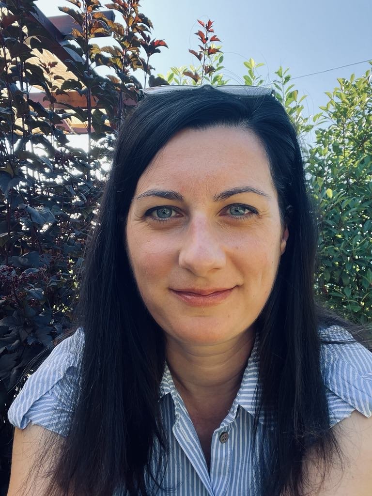 A woman with long dark hair and blue eyes smiling outdoors, wearing a blue and white striped shirt, with trees and clear sky in the background.