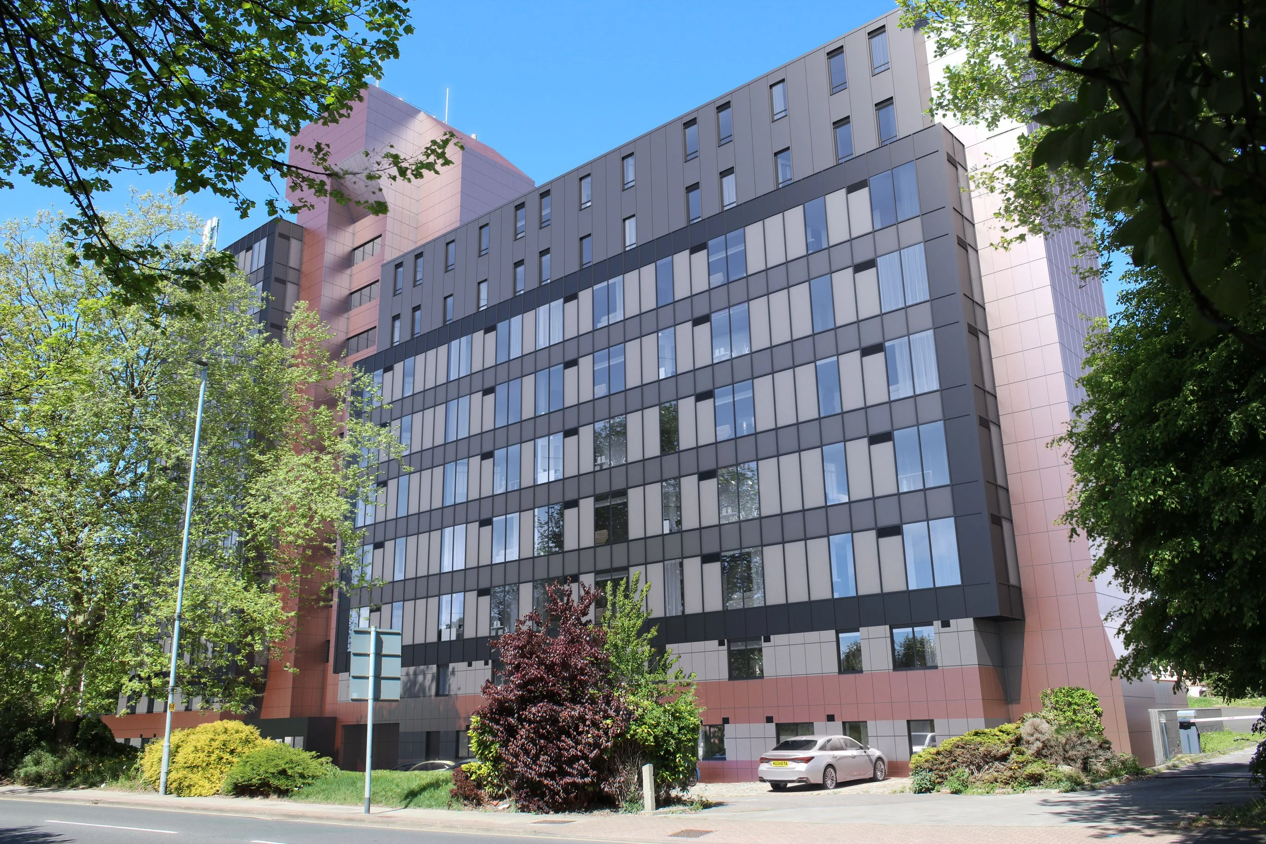 A modern multi-story office building with reflective glass windows, surrounded by green trees and foliage, under a clear blue sky.