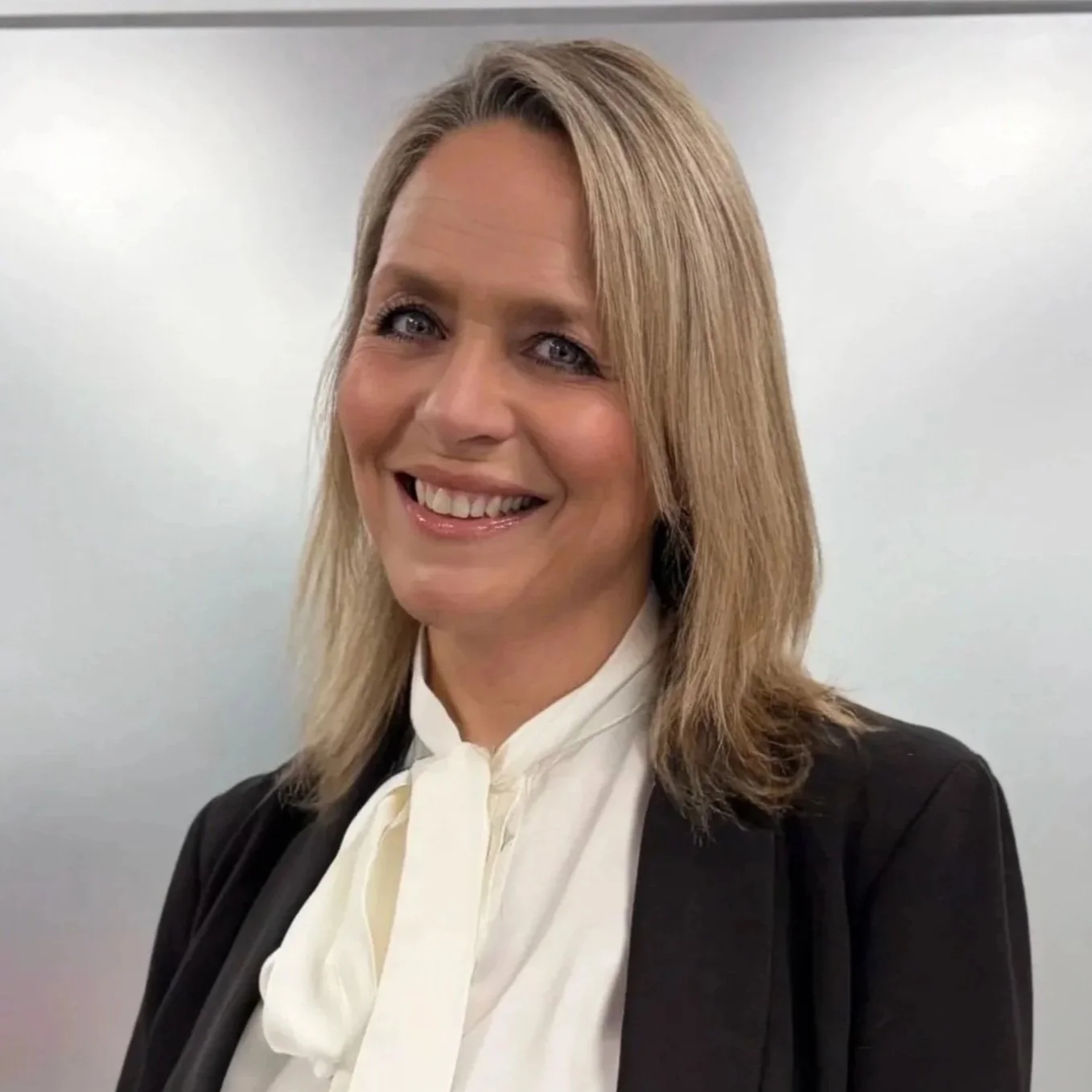 A woman with shoulder-length blonde hair smiling, dressed in a white blouse and dark blazer against a plain neutral background.