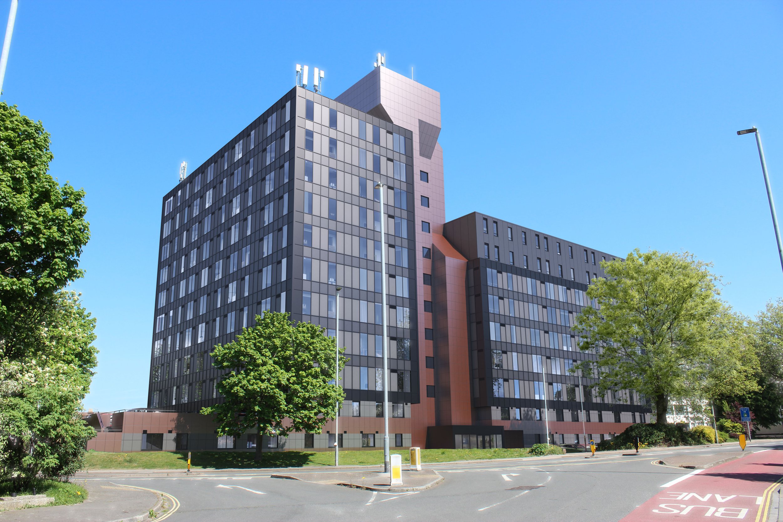 A modern tall building with a black and pink exterior, surrounded by a few trees, streetlights, and a paved road under a clear blue sky.
