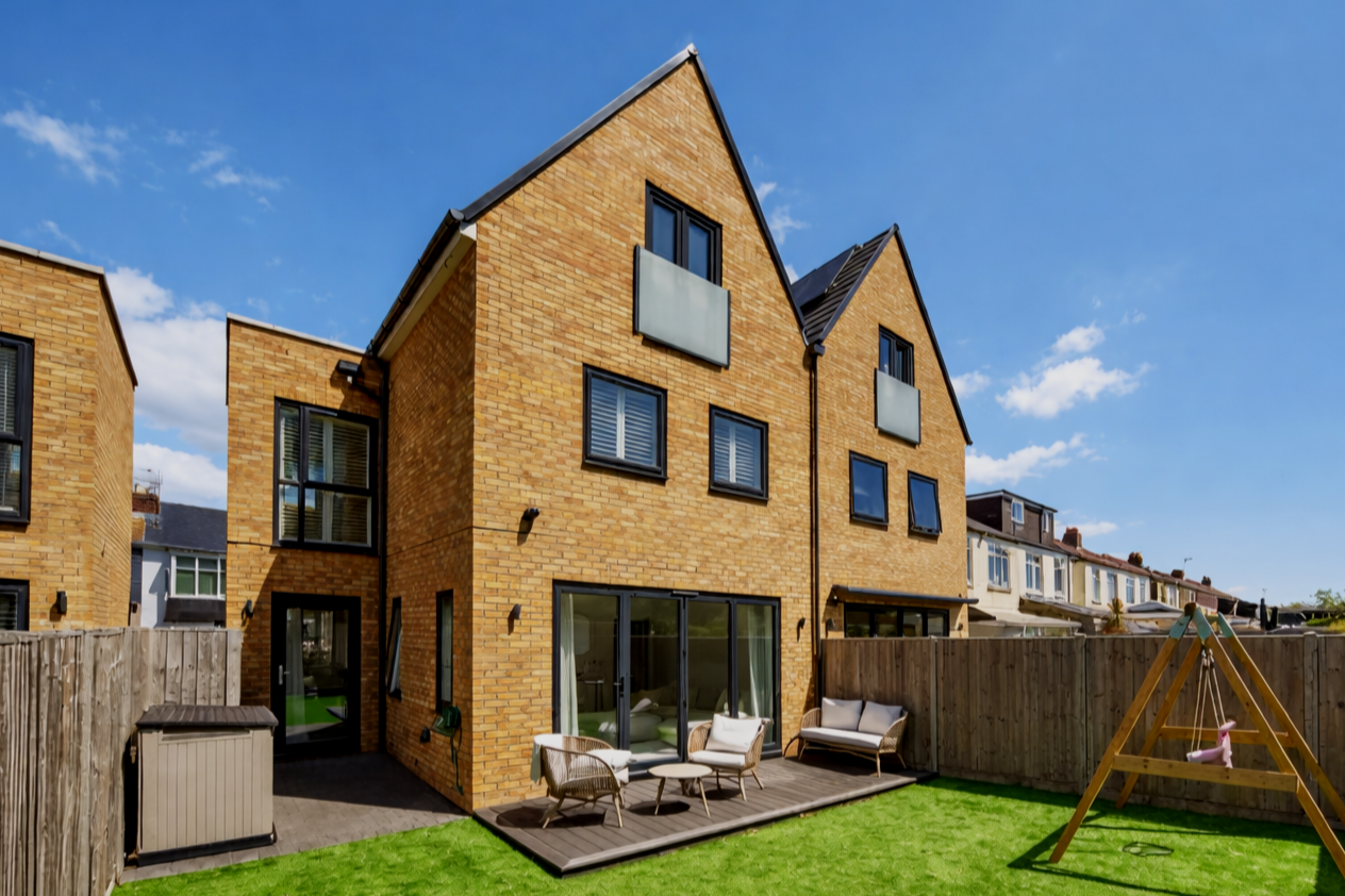 Backyard view of a modern, multi-story brick house with outdoor furniture, a wooden deck, a swing set, and a green lawn under a blue sky with clouds.