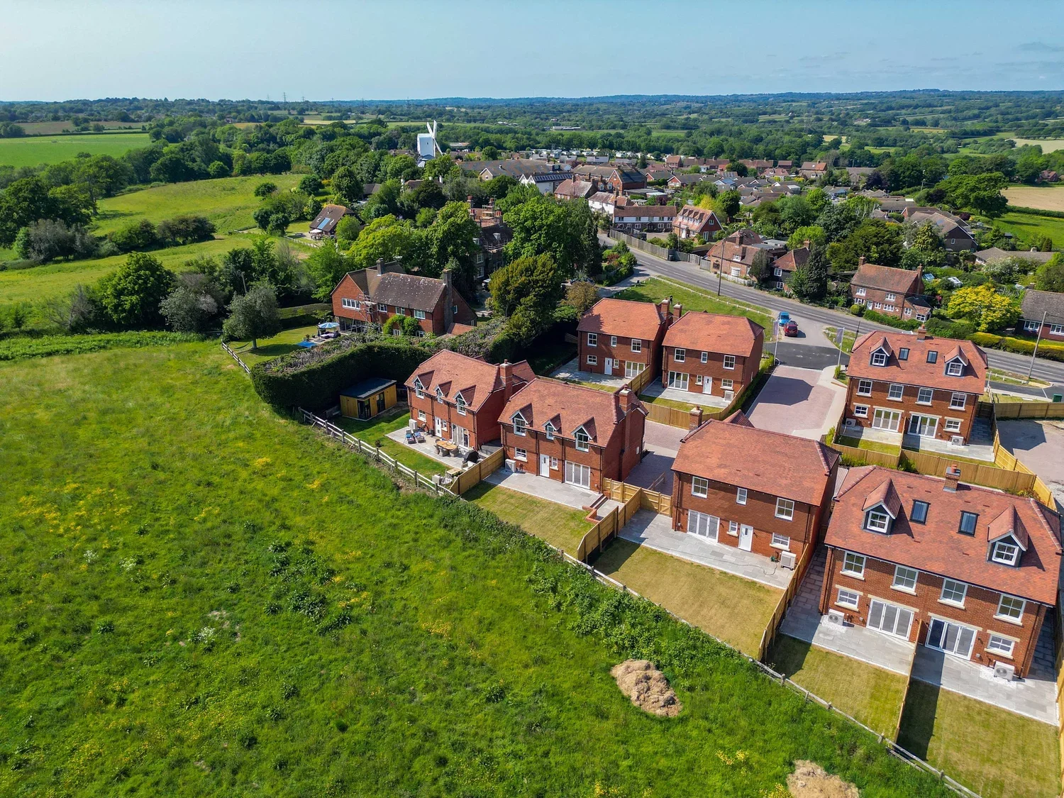 Aerial view of a suburban neighborhood with multiple brick houses, green lawns, and surrounding greenery, with a rural landscape in the background.