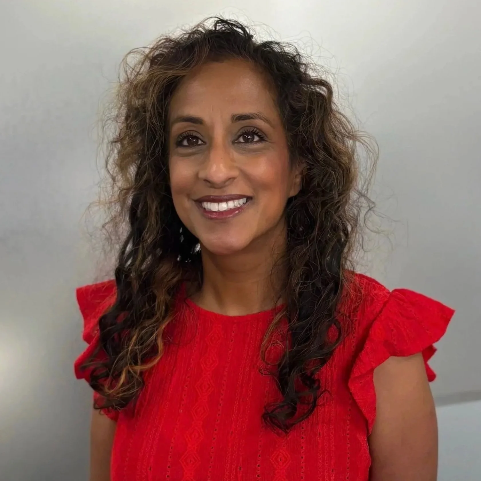 A woman smiling with long, curly hair wearing a red top against a plain background.