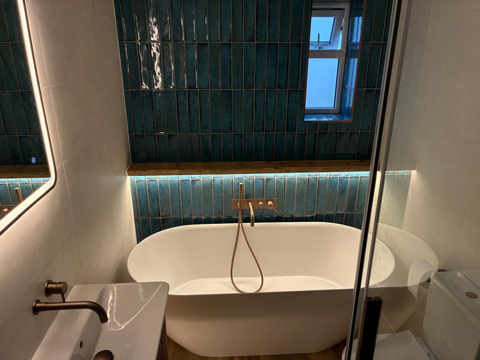 A showroom display featuring travertine brick tiles in a staggered vertical pattern, white suite comprising sink, toilet and bath, luxe walk in shower area and tv! The walls and floor are a matching stone effect in a mid brown/beige