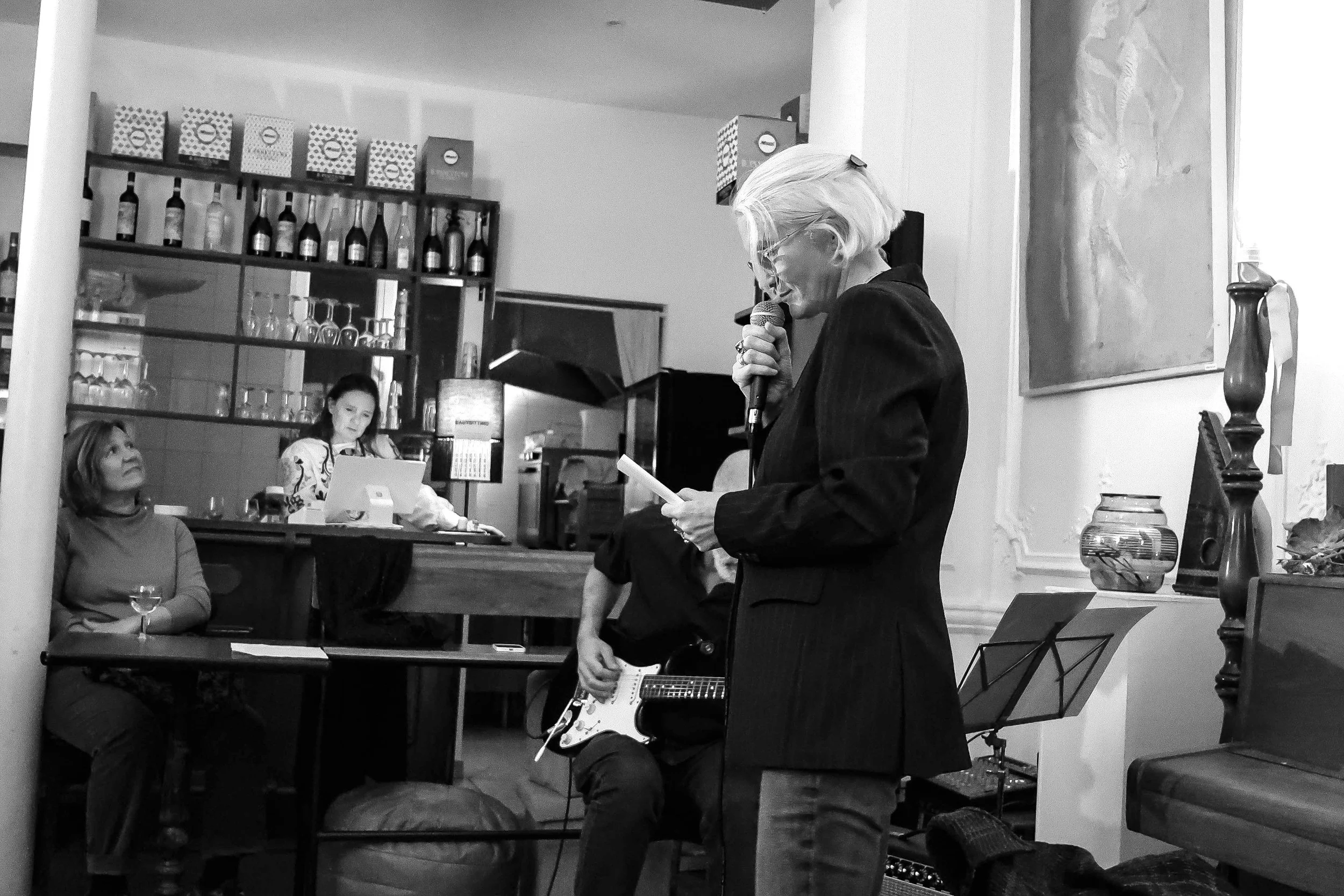 Une femme âgée avec des cheveux blonds courts, portant un costume noir, lit un texte à haute voix avec un microphone dans un café ou un bar, pendant qu'un homme joue de la guitare électrique en arrière-plan, entourée de spectateurs.