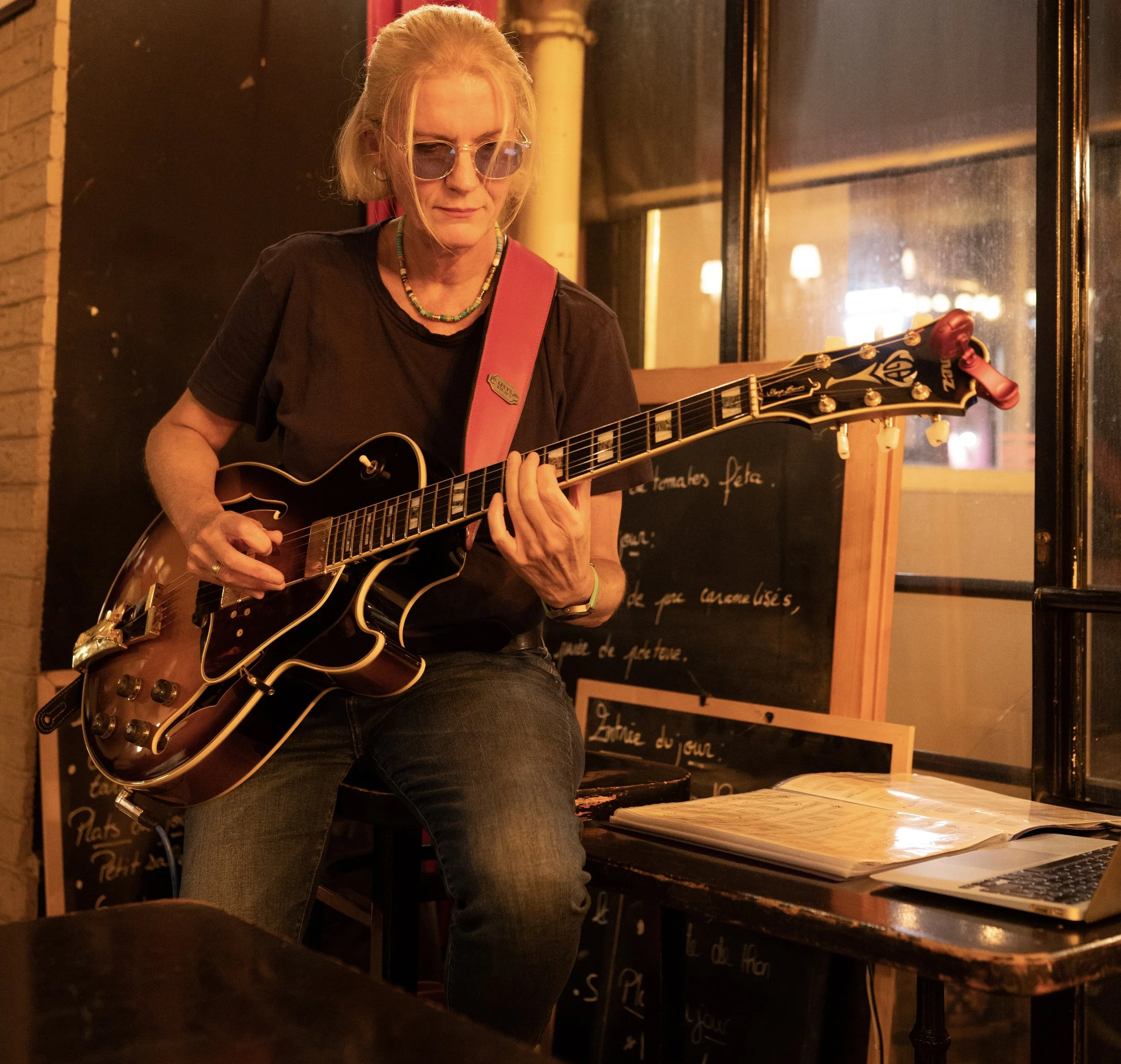 Une femme jouant de la guitare dans un café, avec des notes manuscrites sur un tableau noir en arrière-plan.