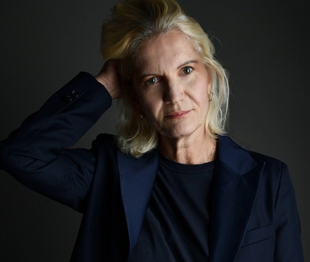 Femme blonde en tenue noire, regardant l'objectif, tenant la tête avec une main, fond sombre.