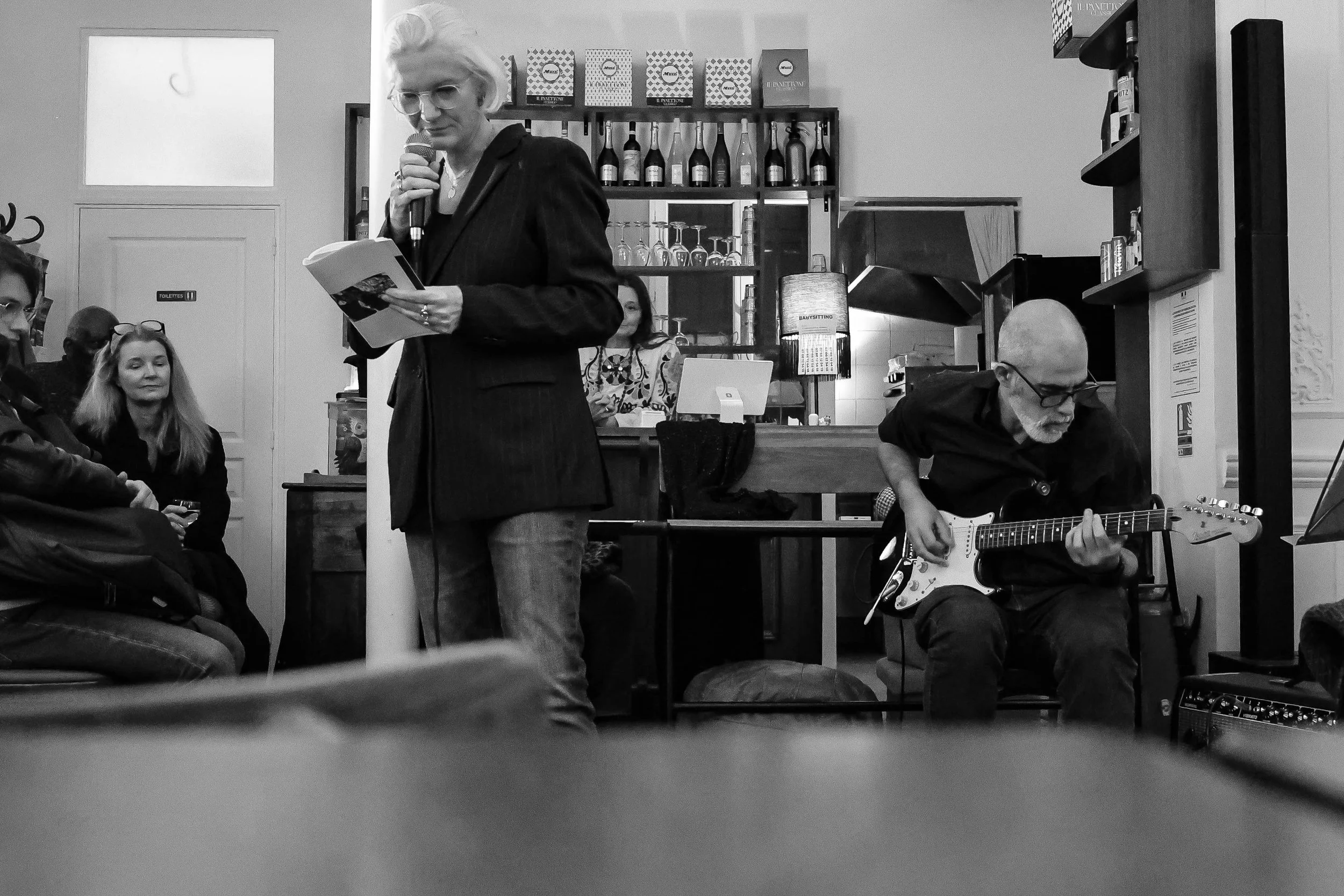 Une femme lit un livre ou un poème devant un public, tandis qu'un homme joue de la guitare électrique dans un café ou un bar, avec plusieurs personnes assises et regardant la scène en arrière-plan, en noir et blanc.