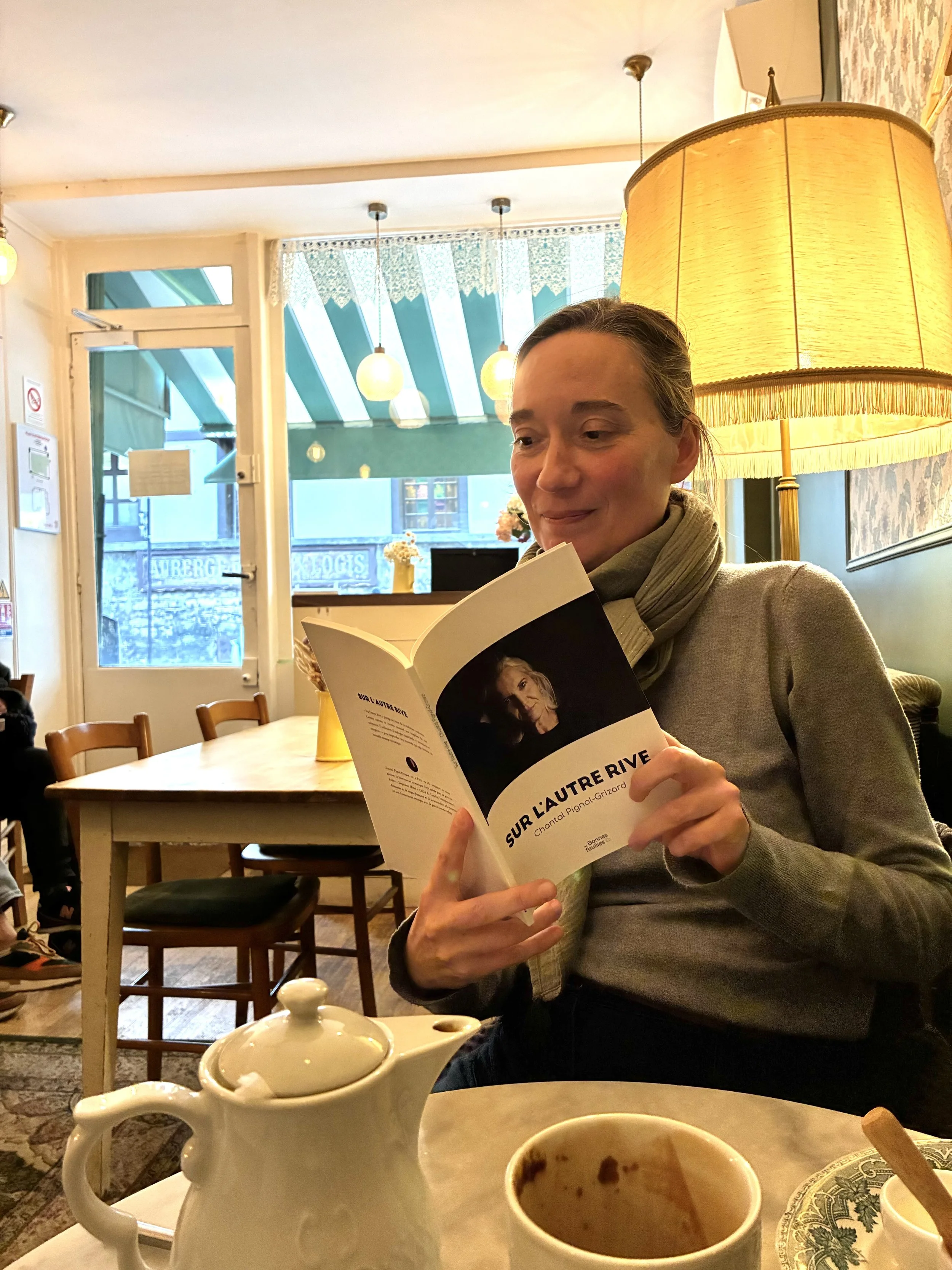 Femme assise dans un café lisant un livre intitulé "Sur l'autre rive" sous une grande lampe.