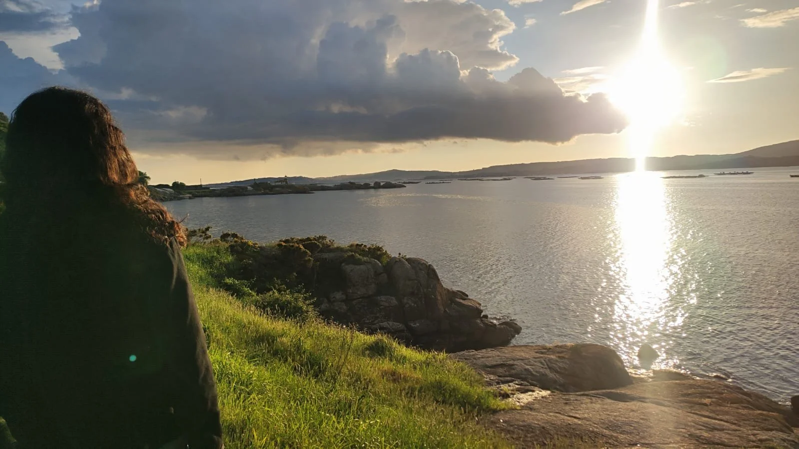 A person with long curly hair sitting on grass and rocks by a body of water, watching the sunset with clouds overhead.