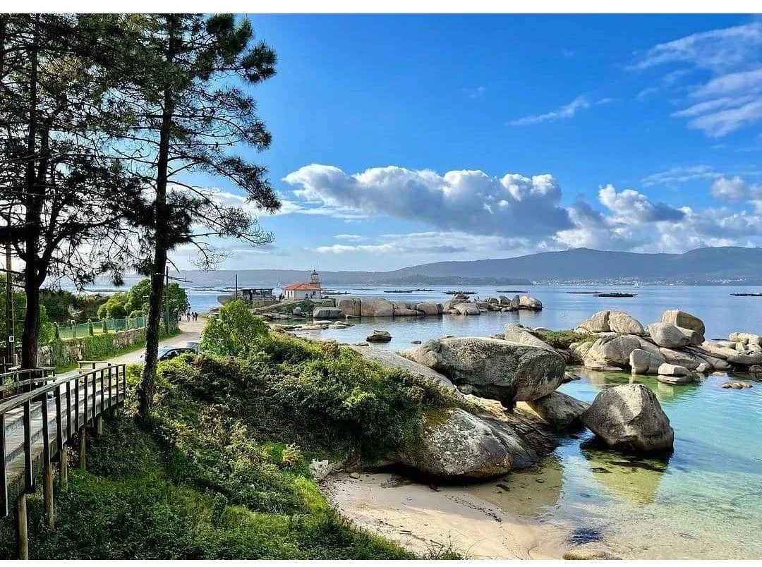 A coastal scene with large rocks along the shore, a sandy beach, lush greenery, tall trees, a fence, a pathway, a small lighthouse, and water with distant mountains and clouds in the sky.