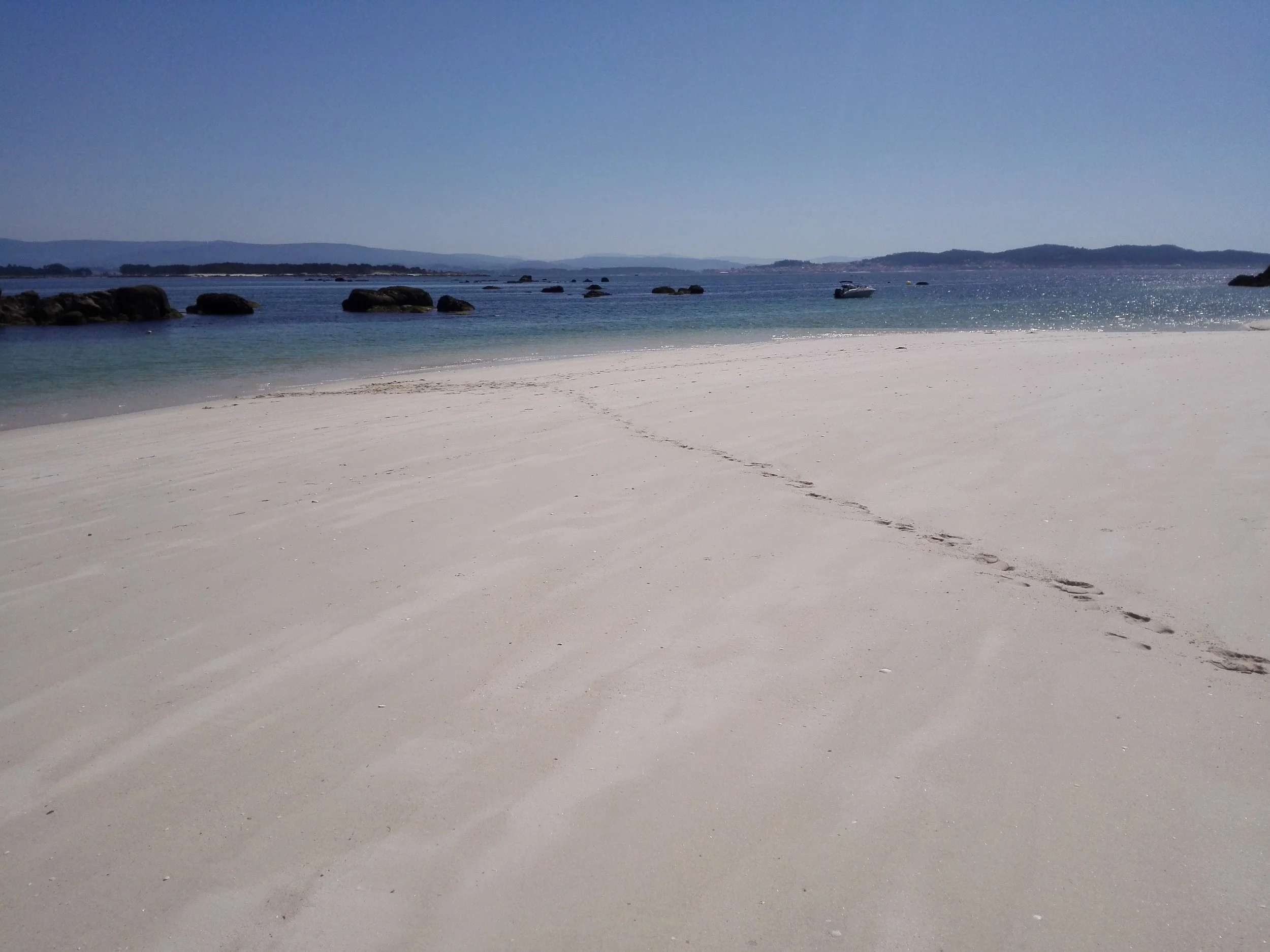 Empty sandy beach with footprints leading toward the water, rocks in the ocean, boat in the distance, and calm blue sky.