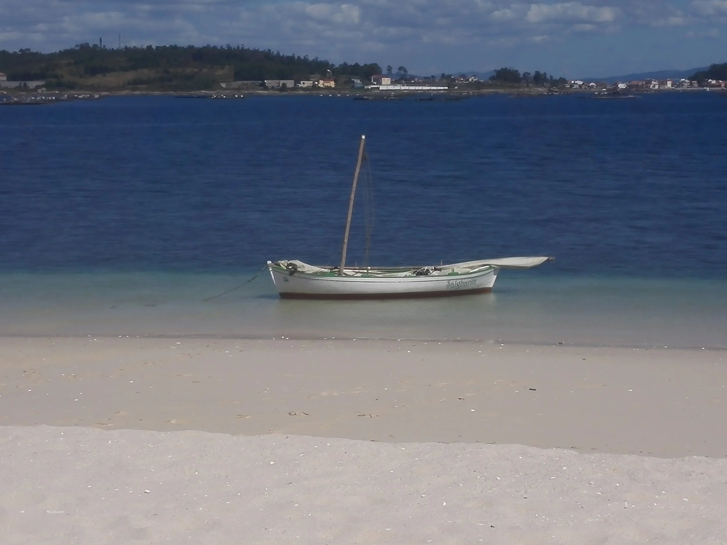 A small sailboat anchored near the shore in calm blue water, with a sandy beach in the foreground and a distant shoreline with buildings and trees in the background.