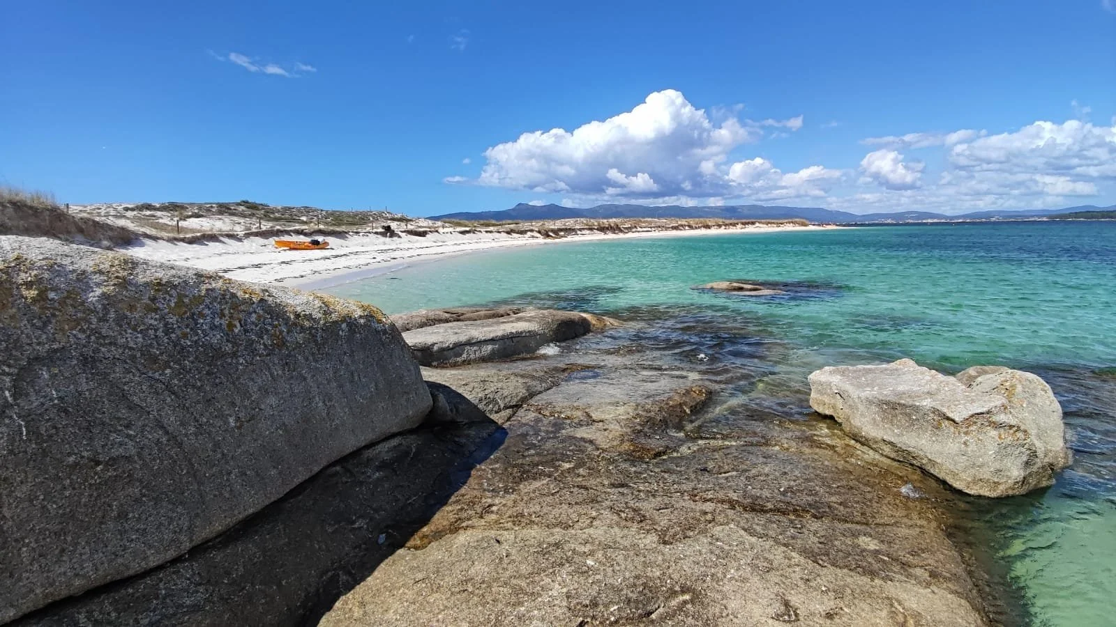 Beach with large rocks near the shoreline, turquoise water, sandy beach, cloudy sky, distant mountains.