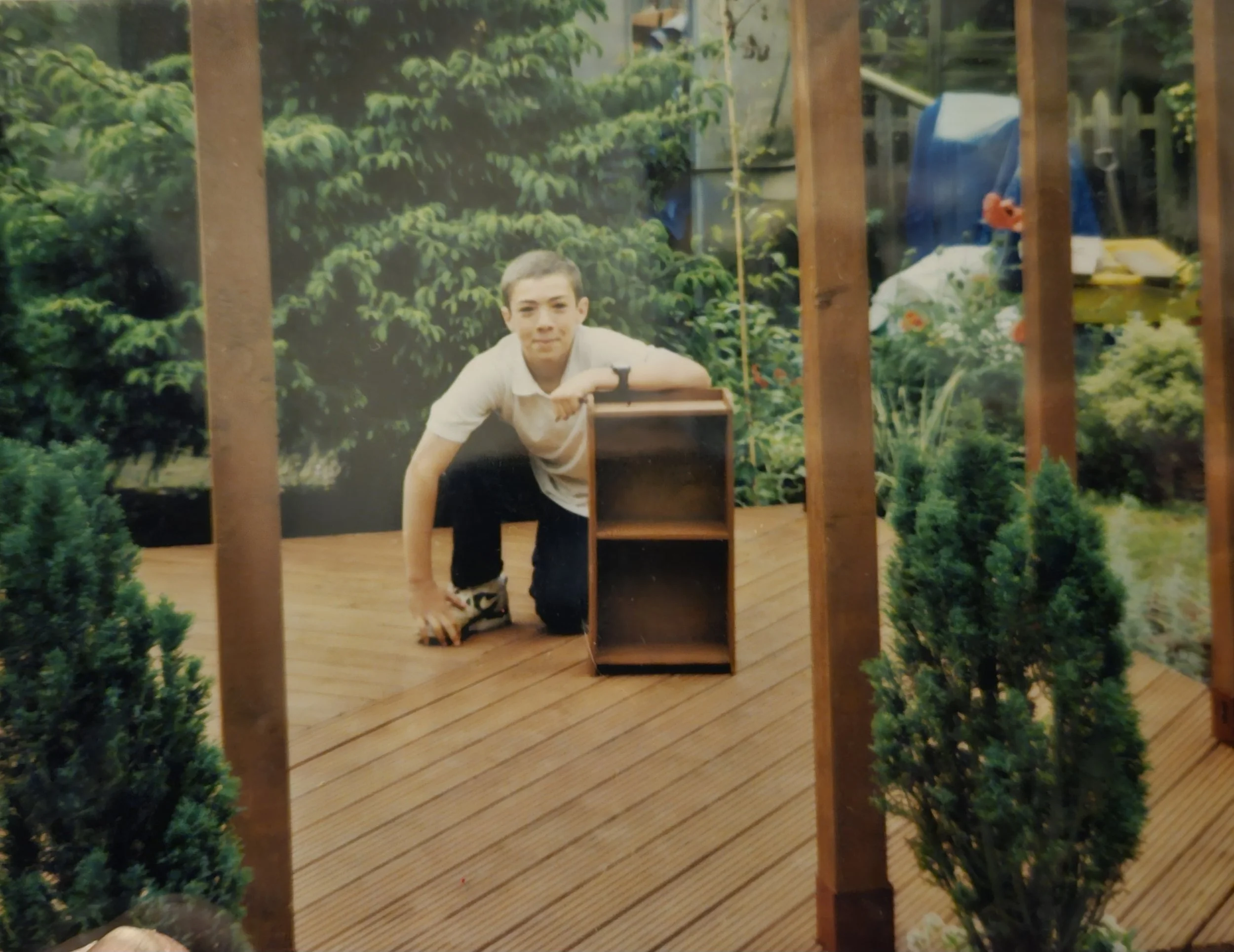 A young boy on a wooden deck outdoors, crouching down with one hand on the deck and the other on a small wooden bookshelf, surrounded by green trees and plants.