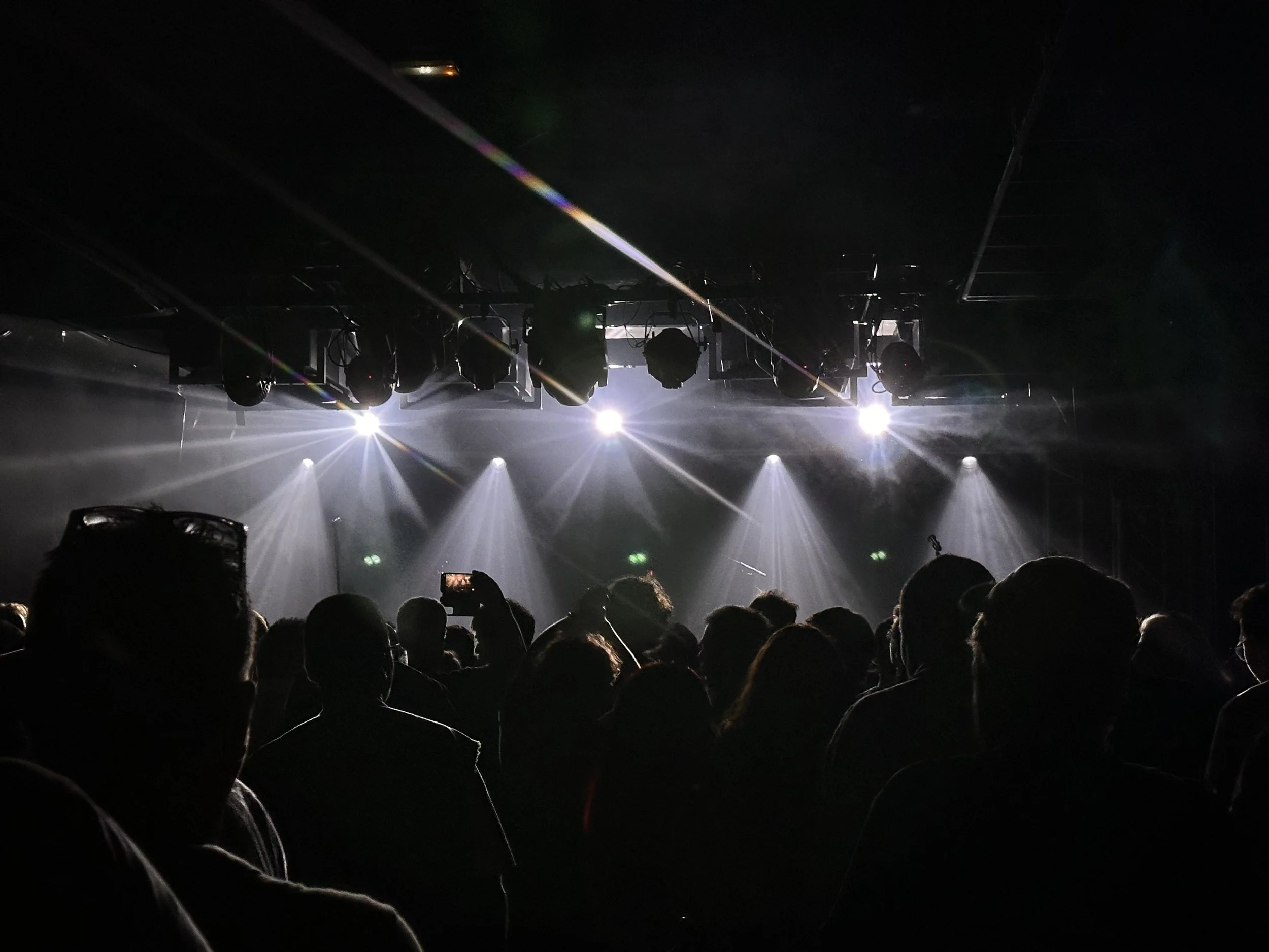 Public assis dans une salle sombre, éclairée par des projecteurs blancs, prête pour un spectacle ou un concert.
