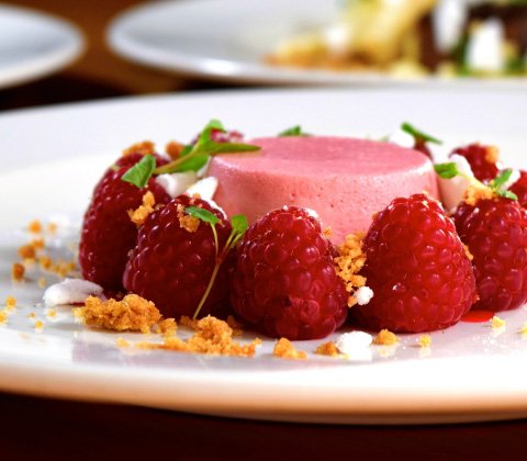 Gourmet dessert with pink mousse, fresh raspberries, and crumble on a white plate.