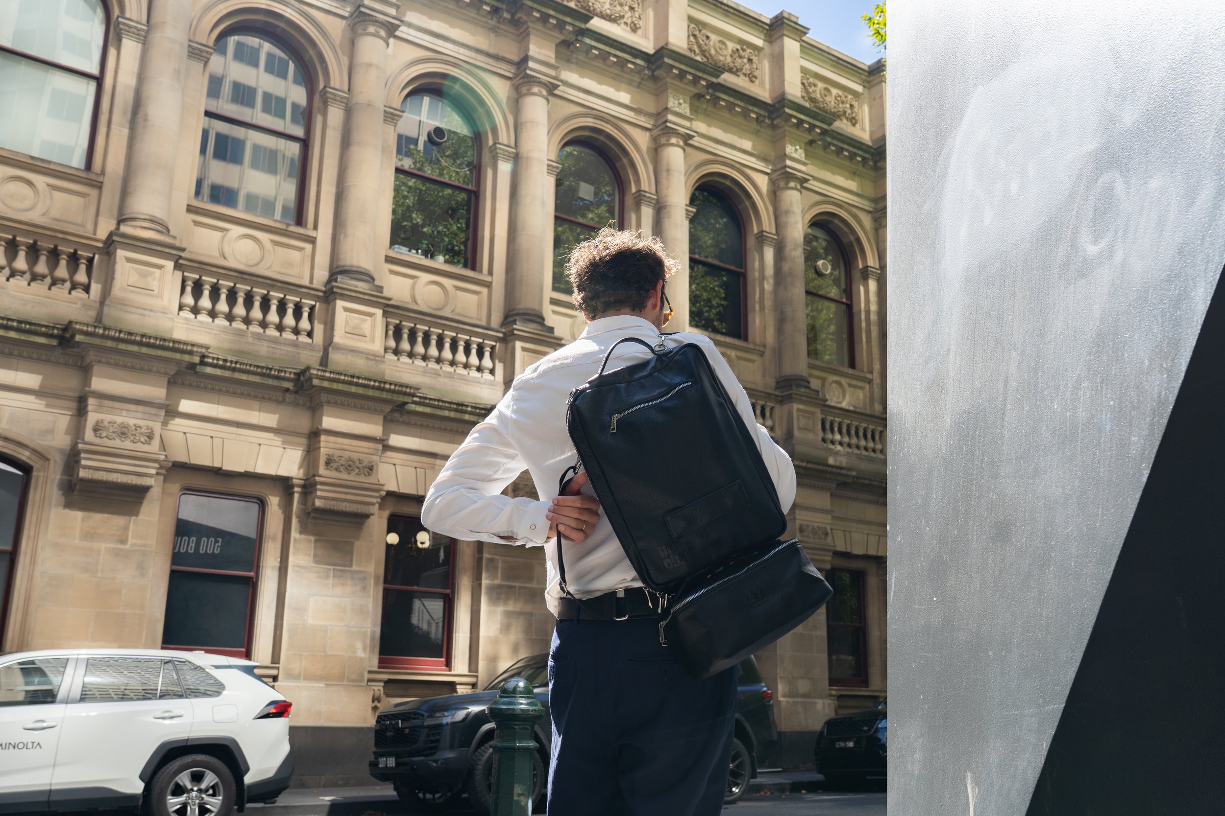 Man with curly hair wearing glasses, a white shirt, and a black backpack standing on a city street, facing a historic stone building with ornate windows and columns, with parked cars in the background.