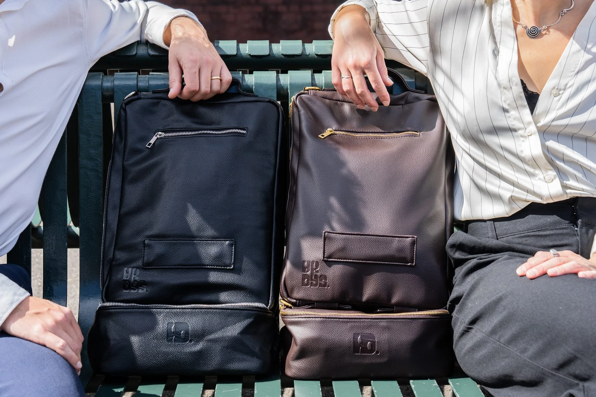 Two people on a bench holding the black and brown WKDYS convertible work bags with detachable lunchboxes