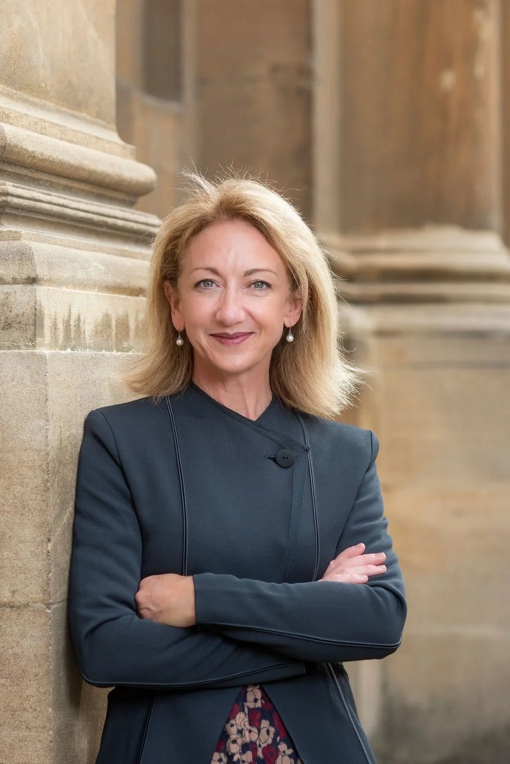 A woman with blonde hair, blue eyes, and pearl earrings, wearing a dark blazer, standing against a stone building with classical architectural features.